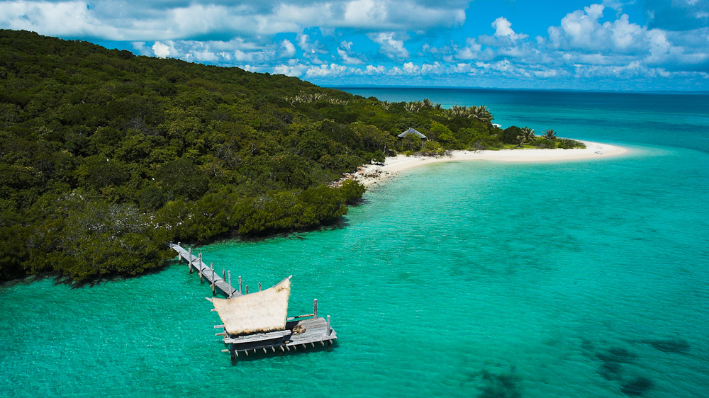 A scenic view of a secluded beach with white sand, surrounded by lush green trees and calm turquoise waters. A wooden dock extends into the water, leading to a small thatched-roof structure. Fluffy white clouds float in a bright blue sky.