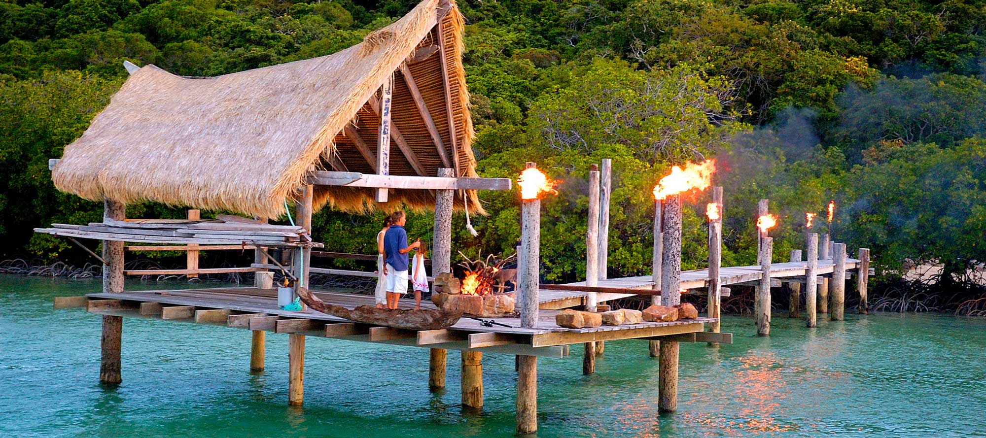 A thatched-roof structure on a wooden dock over turquoise water, surrounded by lush greenery. Torches along the pier are lit, creating a warm atmosphere. Two people are working near the edge of the dock.