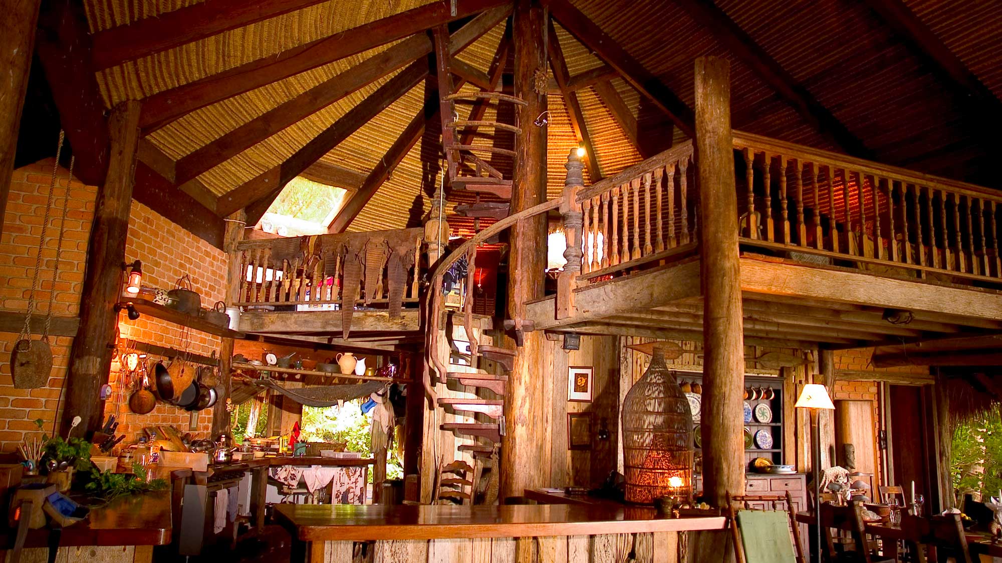 A cozy, rustic cabin interior featuring a spiral staircase, wooden beams, and an open layout. The space includes a kitchen area, seating, and a loft, with warm lighting and decorative items throughout.