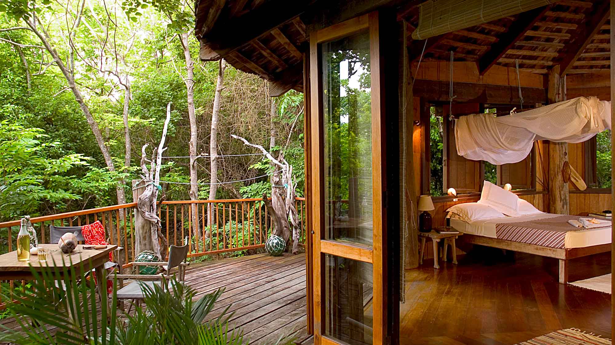 A cozy wooden cabin interior with a bed draped in white fabric, opening to a lush green forest. A small table and chairs are on the deck, surrounded by trees and greenery. Natural light fills the space, creating a warm and inviting atmosphere.