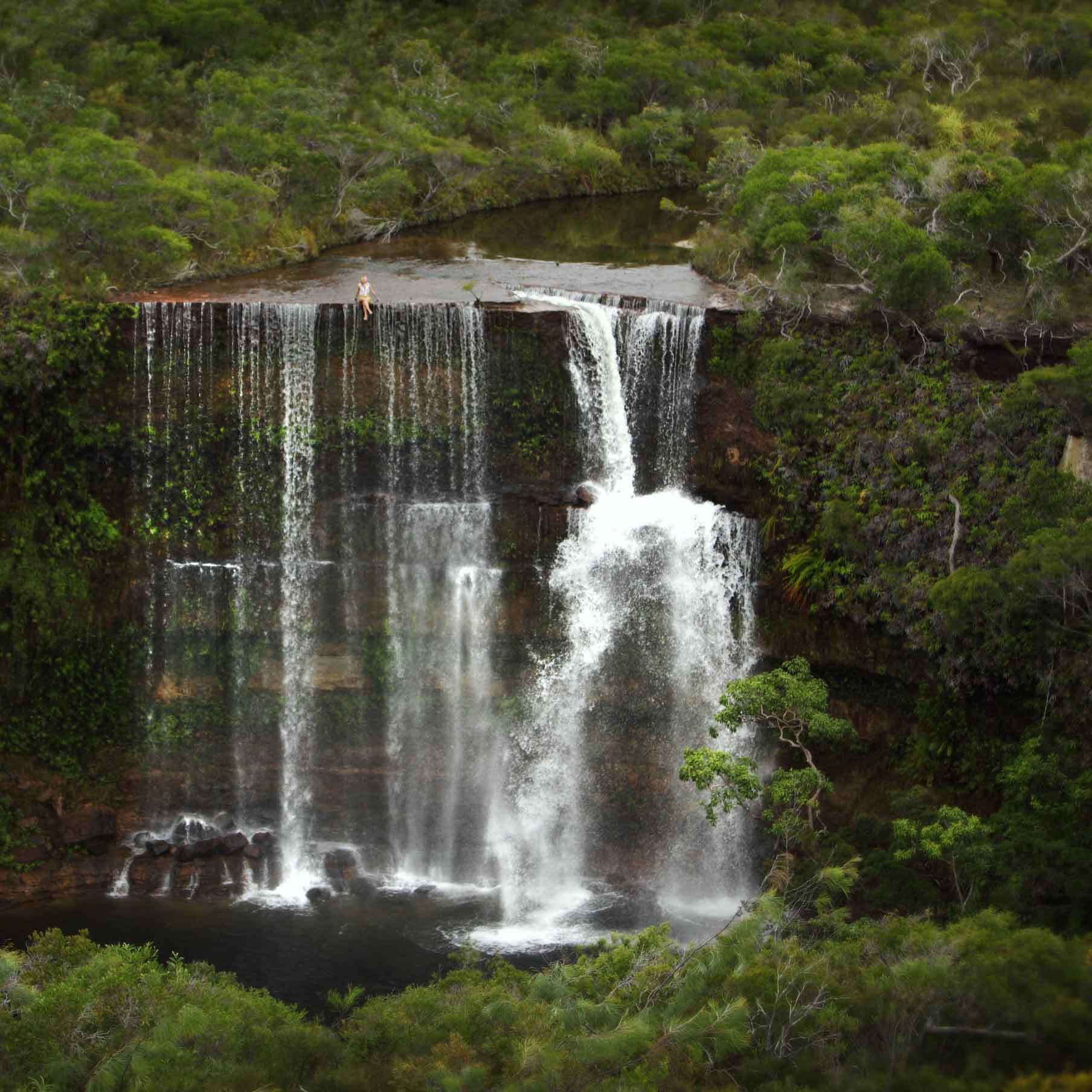 A lush green landscape featuring a dramatic waterfall cascading over rocky cliffs into a serene pool below. The surrounding area is dense with vegetation, highlighting the natural beauty of the scene.