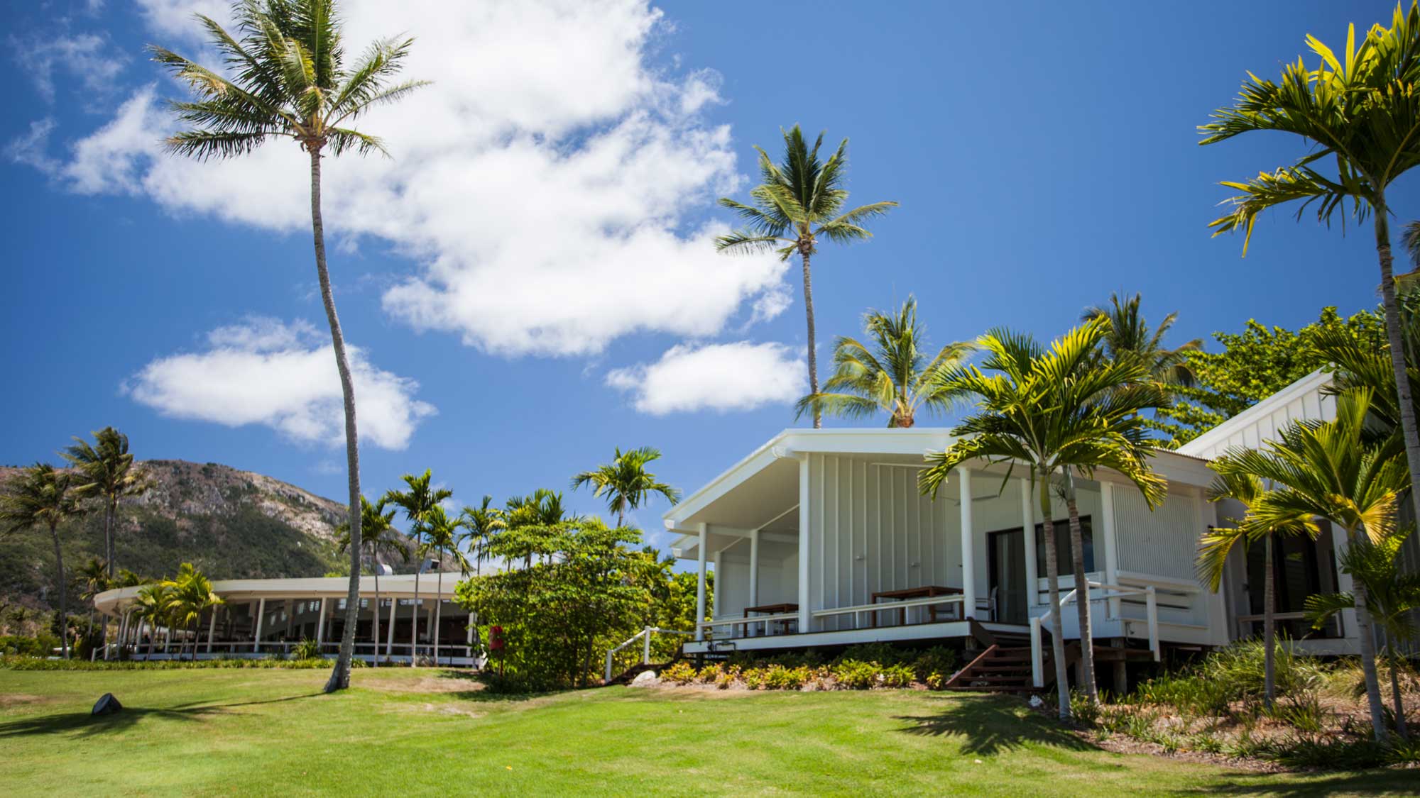 Tropical resort with modern bungalows surrounded by palm trees and lush green grass under a bright blue sky with fluffy clouds.