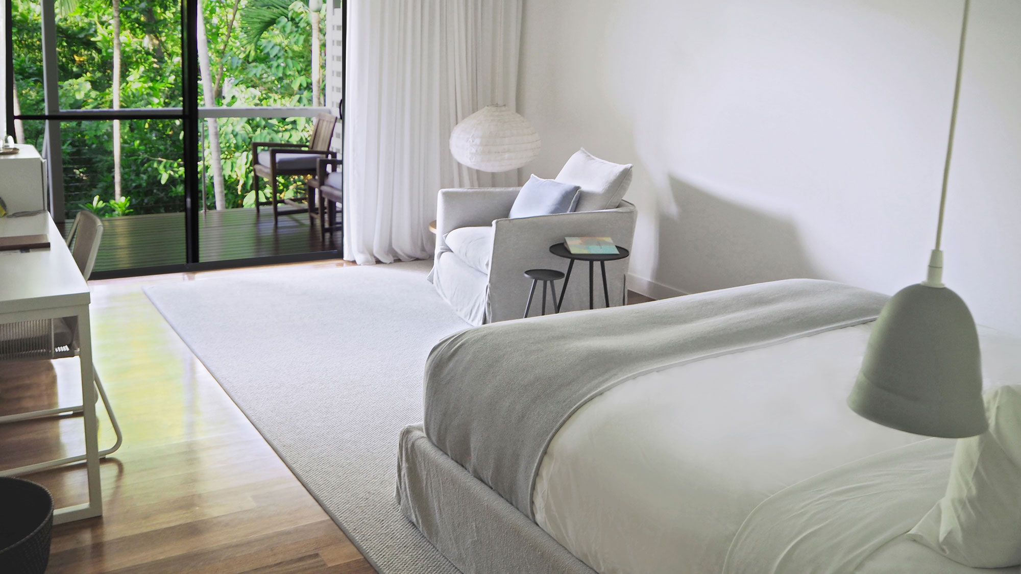 A modern, minimalistic bedroom featuring a large bed with a grey throw, a comfortable armchair, and a small side table. Large glass doors lead to a balcony surrounded by greenery. A desk and chair are positioned nearby on wooden flooring.