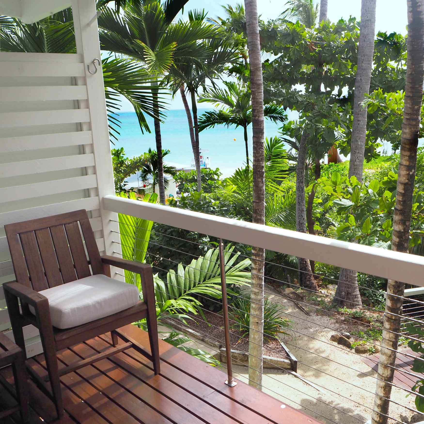 A wooden chair with a cushion sits on a balcony overlooking lush tropical greenery and the ocean in the distance. Palm trees frame the view, creating a serene and inviting atmosphere.