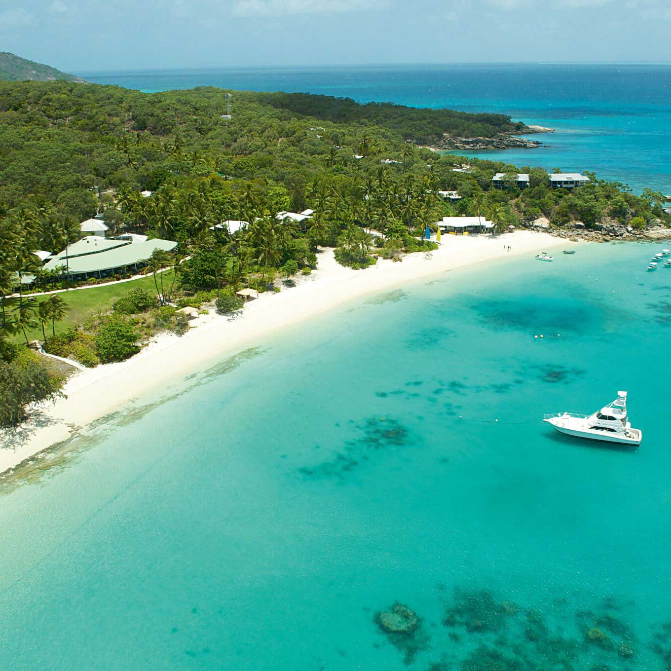 Aerial view of a serene beach with white sand, clear turquoise waters, and lush green vegetation. Small boats are anchored offshore, and tropical palm trees line the coast. The scene evokes a peaceful and sunny tropical paradise.