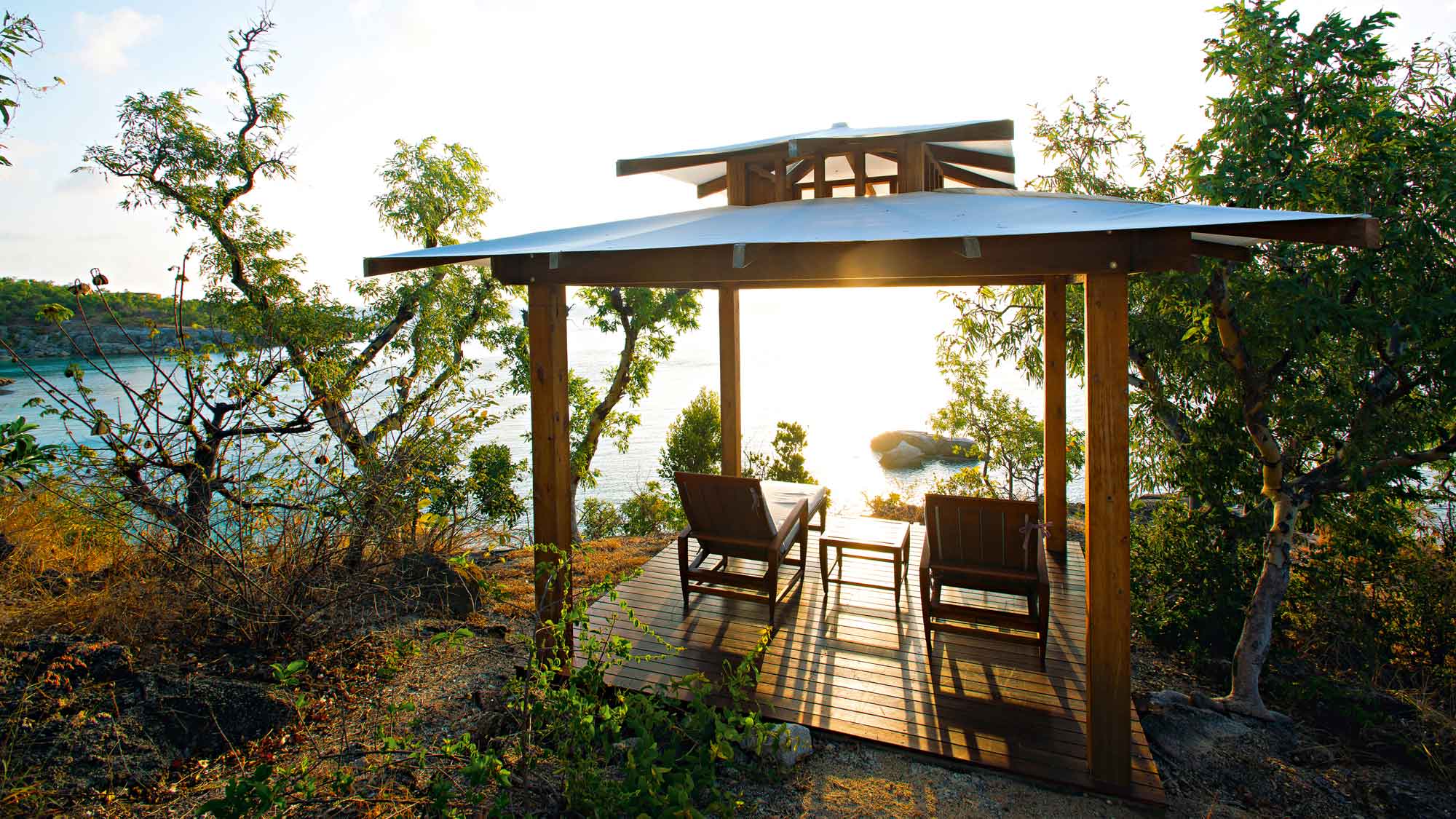 A serene wooden gazebo overlooks a tranquil sea at sunset, featuring two comfortable chairs positioned to enjoy the view. Lush greenery surrounds the area, enhancing the peaceful atmosphere.