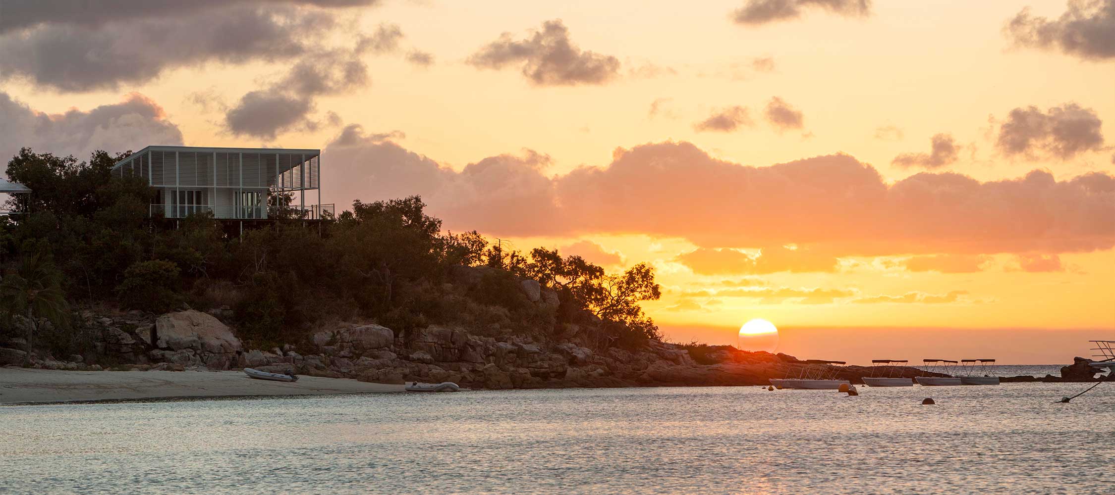 A modern house perched on a hill overlooks a tranquil bay at sunset, with vibrant orange and pink hues in the sky and a sun setting near the horizon. Rock formations and trees frame the scene, creating a serene coastal atmosphere.