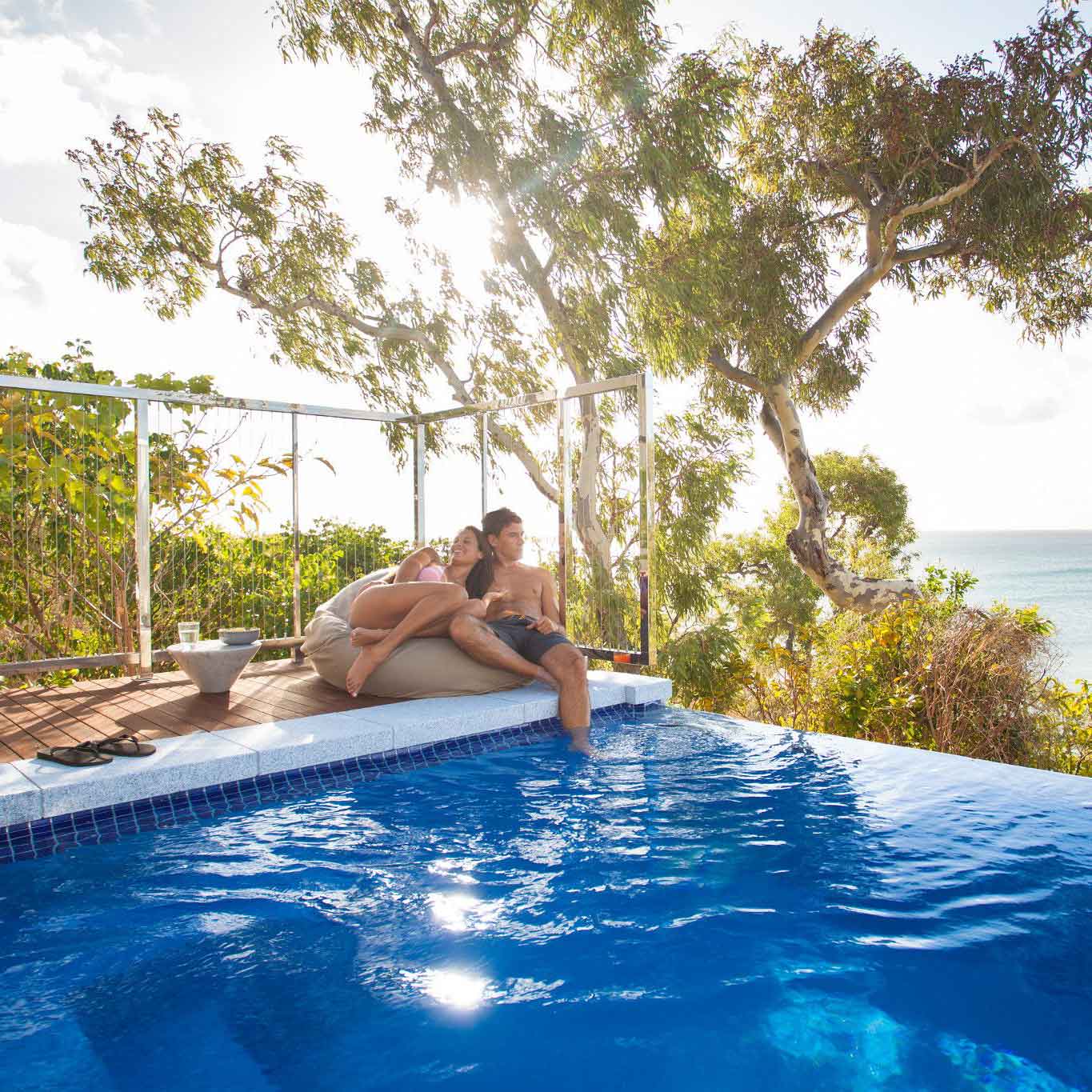 A couple relaxes by a poolside, leaning against each other with a scenic view of the ocean and trees in the background. The sun is shining brightly, creating a warm and inviting atmosphere.