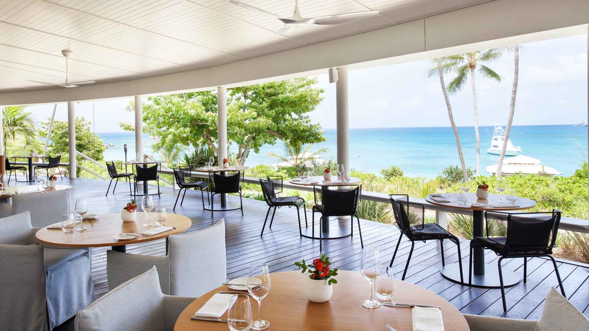 An elegant restaurant interior with wooden tables set for dining, featuring minimalistic decor. The expansive windows offer stunning views of a blue ocean and palm trees, with a gentle breeze suggested by the ceiling fans.