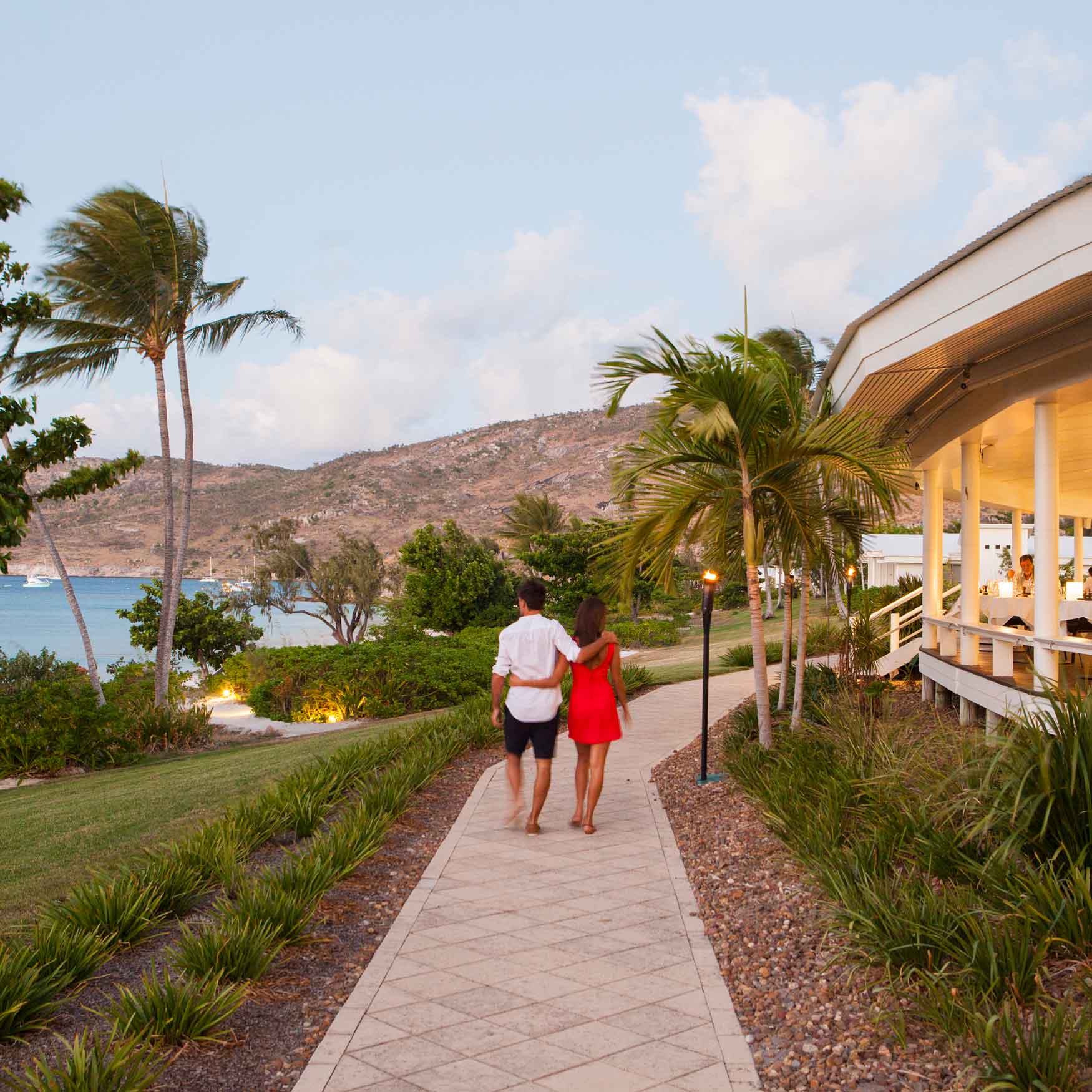 A couple walks hand in hand along a scenic pathway lined with palm trees, leading to a coastal area. The woman wears a red dress, while the man is in a white shirt and shorts. In the background, there are boats on the water and a hillside with greenery.
