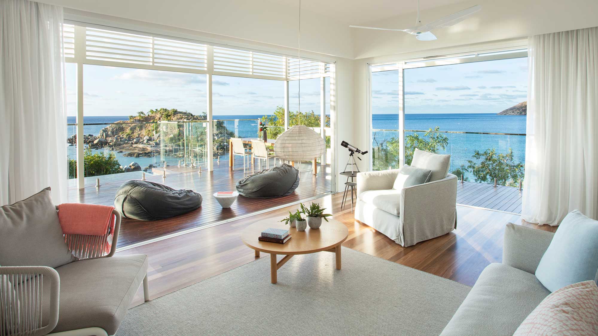 A bright and airy living room with large windows showcasing a stunning coastal view. The space features comfortable seating, a wooden coffee table, and bean bags, with a telescope on the deck for enjoying the scenery. Sunlight floods in, highlighting the natural beauty outside.