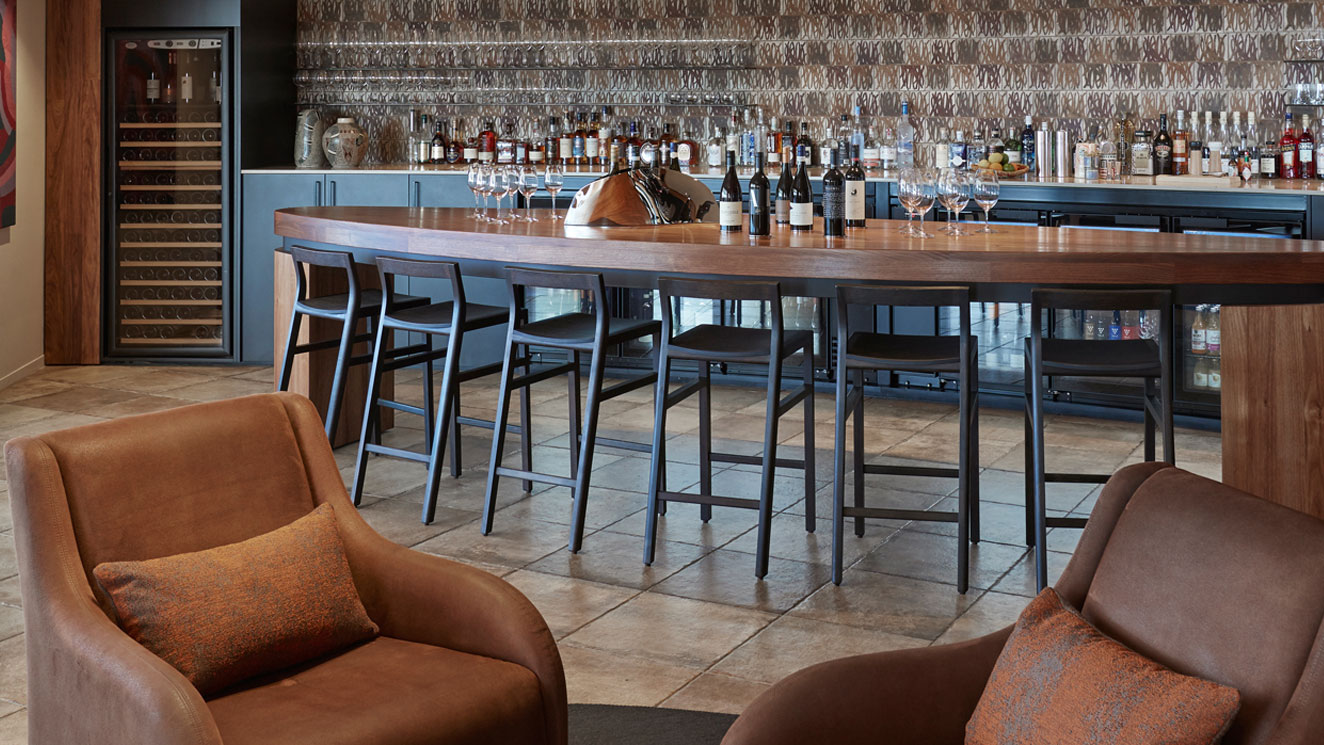 A modern bar with a wooden countertop and high black stools. Behind the bar, shelves display various bottles of liquor. Two brown upholstered chairs with orange cushions are positioned nearby on a tiled floor.