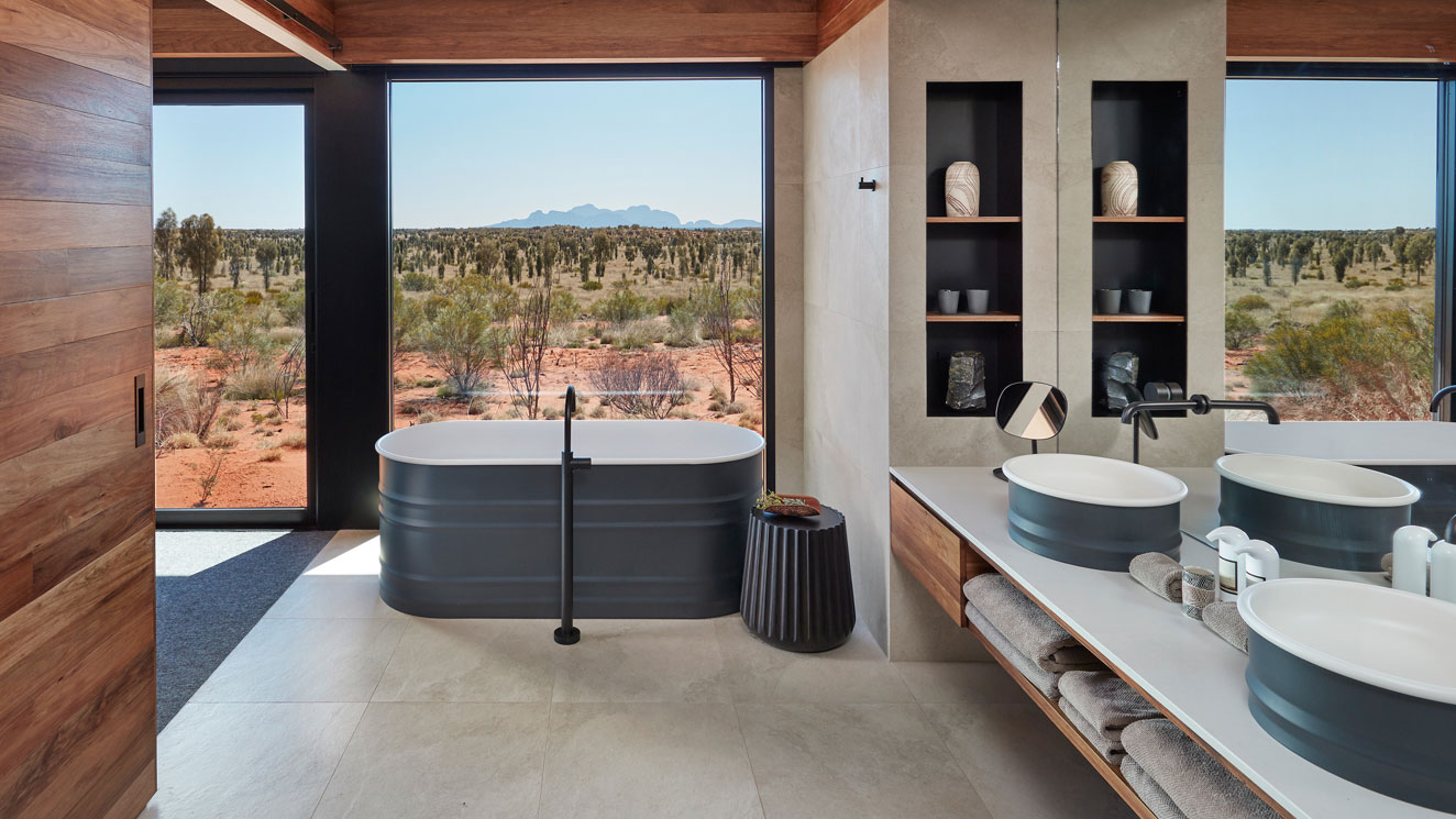 A modern bathroom featuring a freestanding bathtub, double sinks with sleek mirrors, and a view of a desert landscape through large windows. Natural wooden accents and minimalist decor enhance the space.