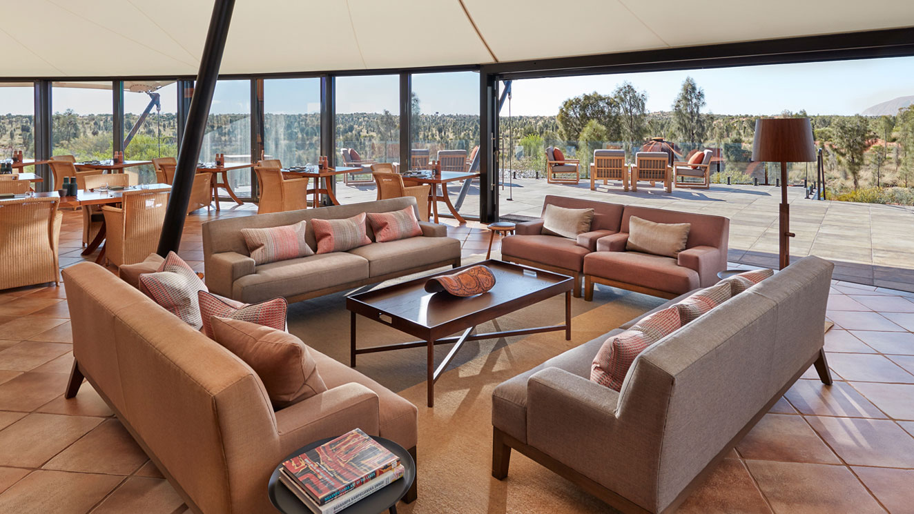 Modern lounge area with beige sofas and a coffee table, surrounded by large windows offering a view of nature. Stylish decor and warm lighting create a welcoming atmosphere.