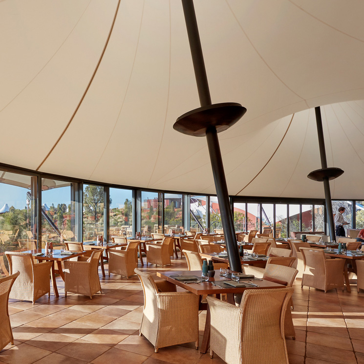 A modern restaurant interior featuring woven wicker dining chairs and wooden tables. The bright space is designed with large glass windows, allowing natural light to flood in, and features a high, sail-like ceiling.