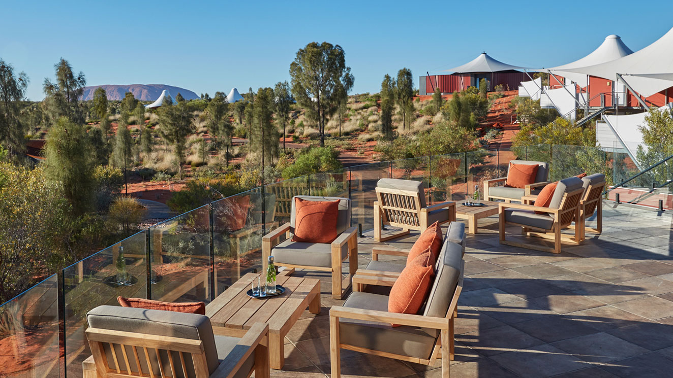 A sunlit terrace featuring modern wooden seating with orange cushions, overlooking a scenic landscape of red earth and sparse vegetation. In the distance, a range of mountains is visible under a clear blue sky.