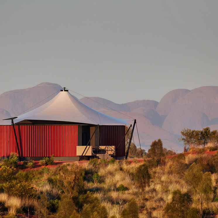 A modern, red and white tented structure sits in a rugged desert landscape, with distant mountains in the background under a soft, warm light.