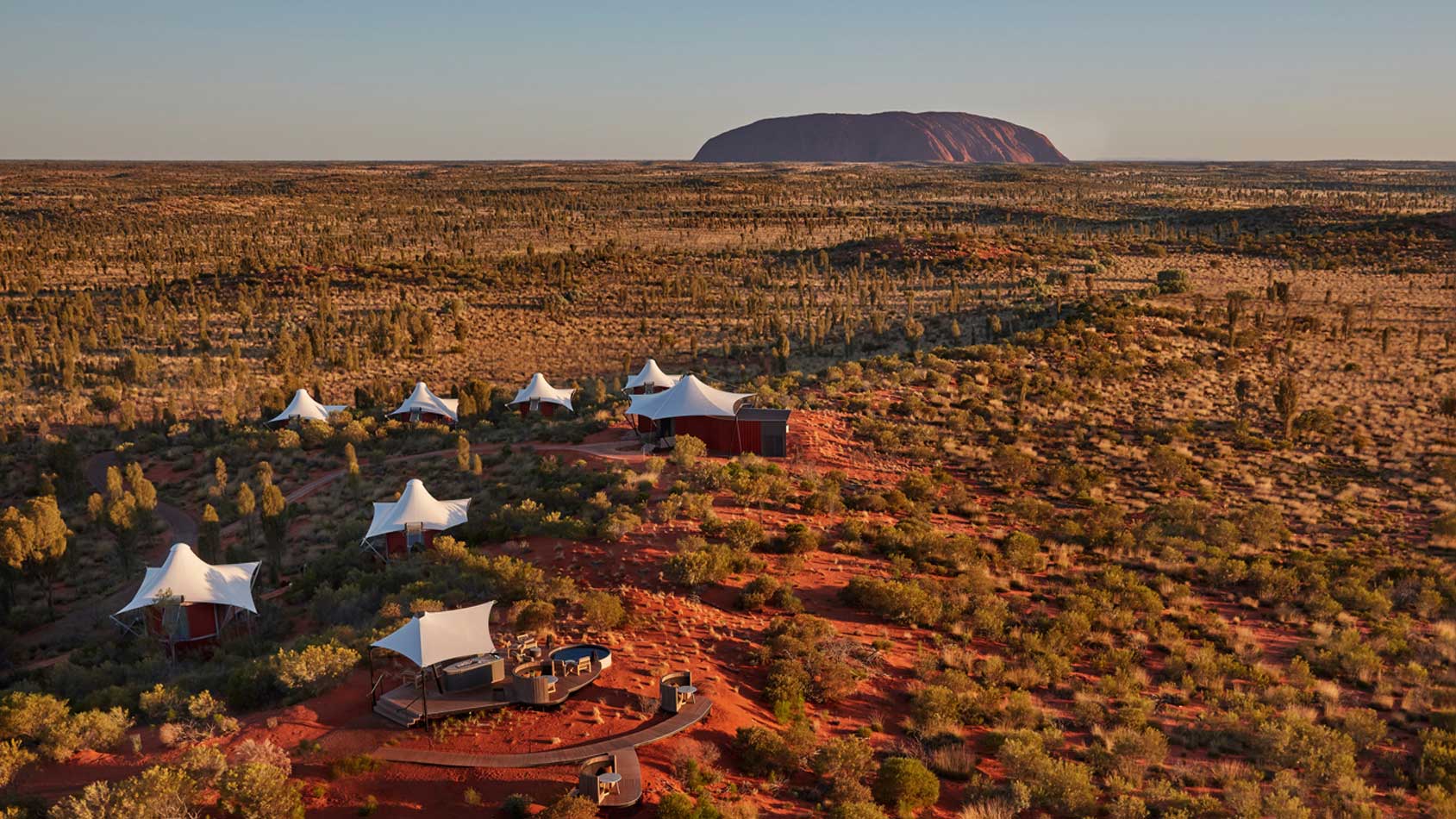 Aerial view of a desert landscape featuring several tent-like structures and a remote outdoor seating area, with the iconic Uluru rock formation in the background, under a clear sky.