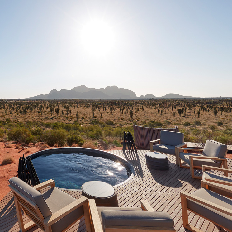 A stylish outdoor seating area with wooden furniture overlooks a vast desert landscape and distant mountains, under a bright sun. A small, circular plunge pool adds a touch of luxury to the serene setting.
