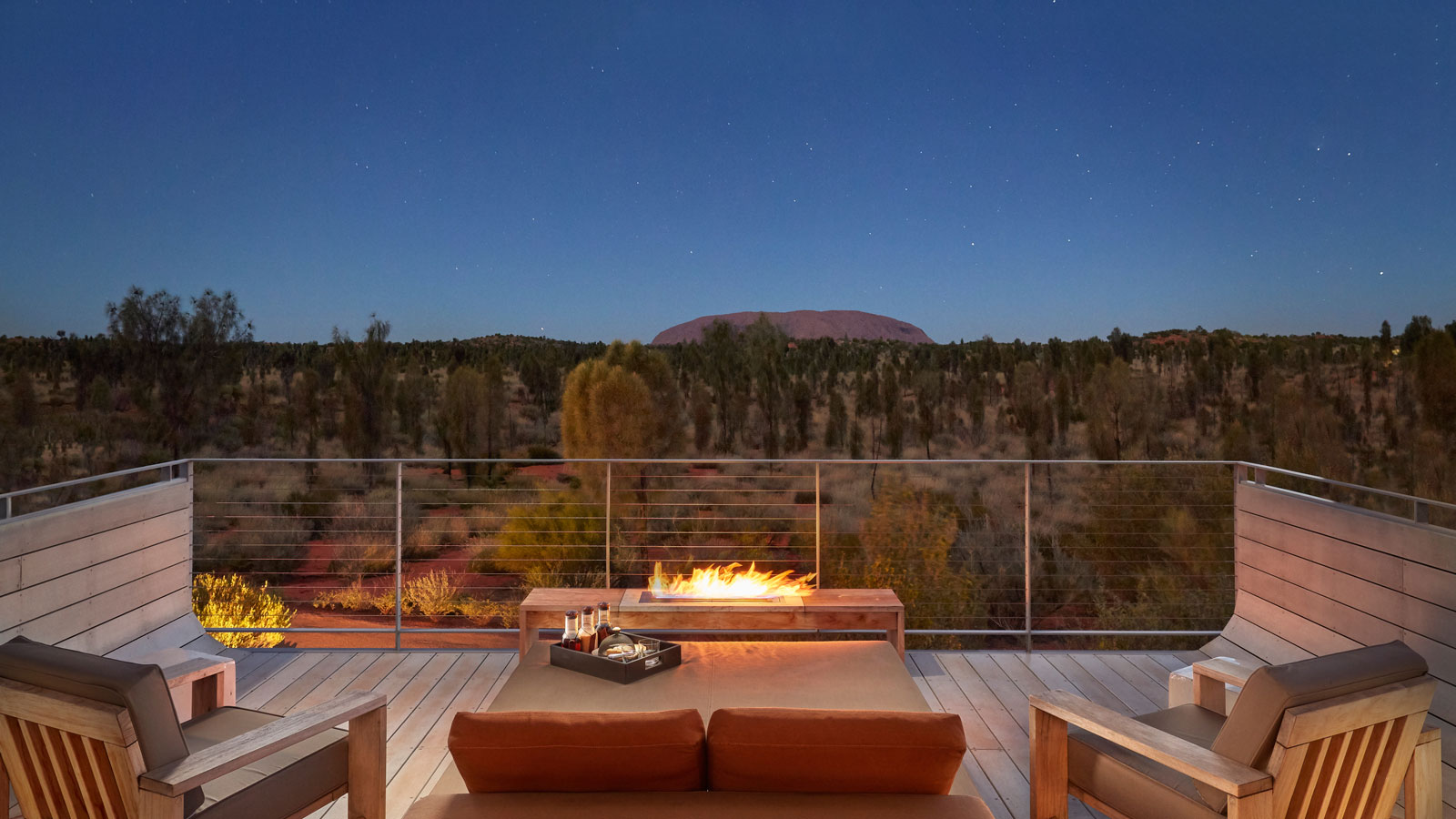 A cozy outdoor seating area with a fire pit, overlooking a vast desert landscape under a starry sky, featuring Uluru in the background.