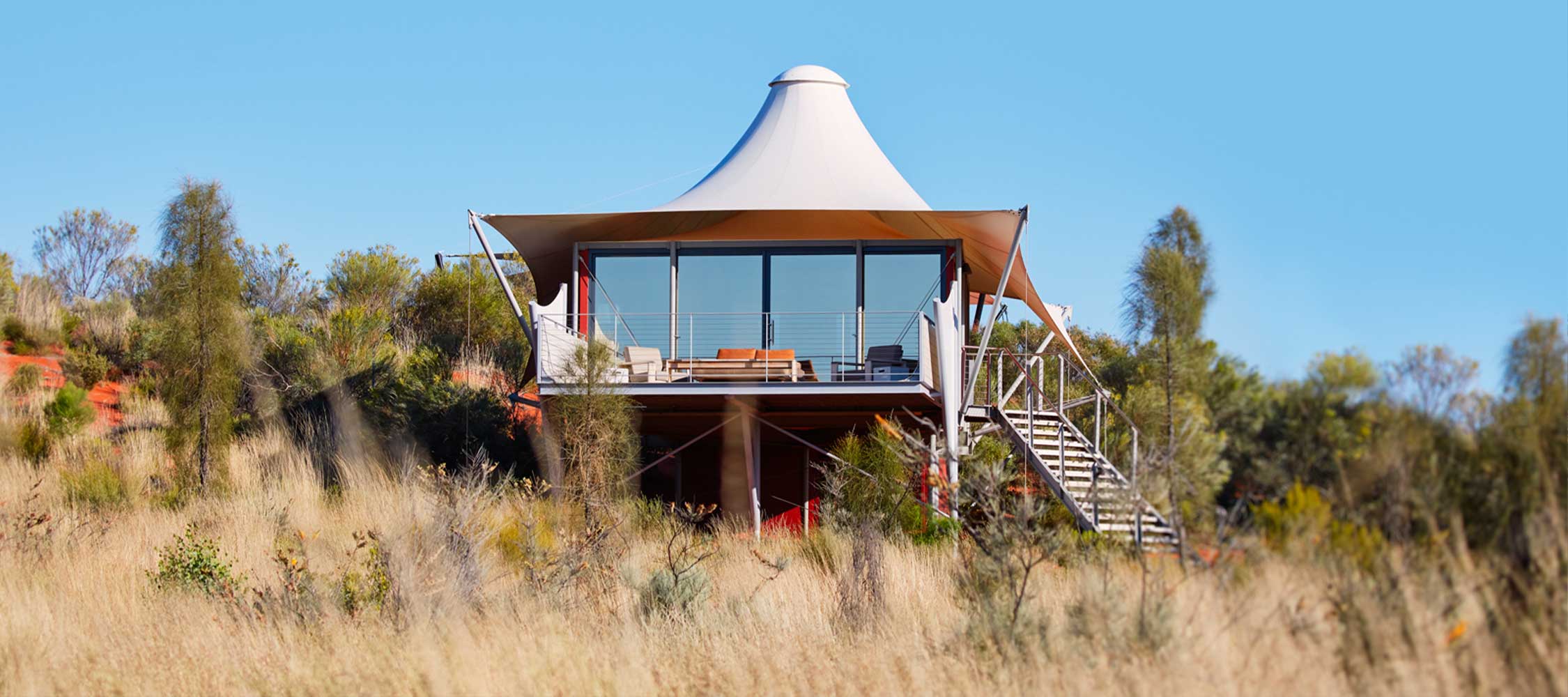 A modern, tent-like structure sits on a hillside surrounded by tall grasses and shrubs. The building features large windows, a sleek design, and a staircase leading to the entrance. A clear blue sky is visible in the background.