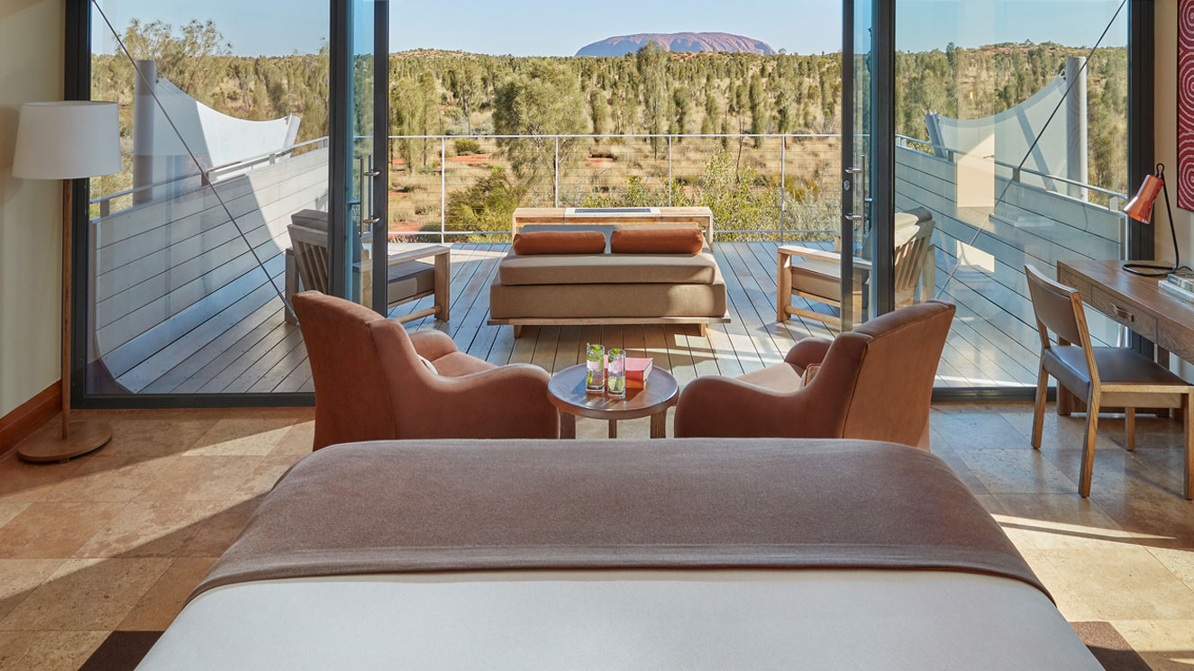 A luxurious hotel room with a king-sized bed in the foreground, two comfortable armchairs, a small round table with drinks, and large glass doors opening to a balcony. The view features a scenic landscape with trees and a distant rock formation, likely Uluru, under a clear blue sky.