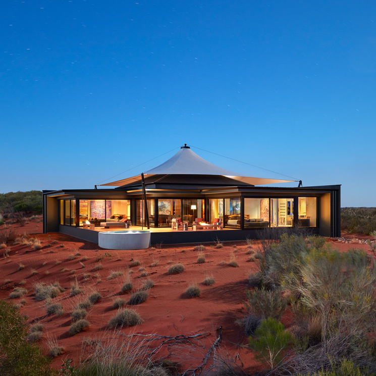 Modern house with a tent-like structure set in a red desert landscape, featuring large glass windows and a circular design. Twilight sky enhances the serene atmosphere.