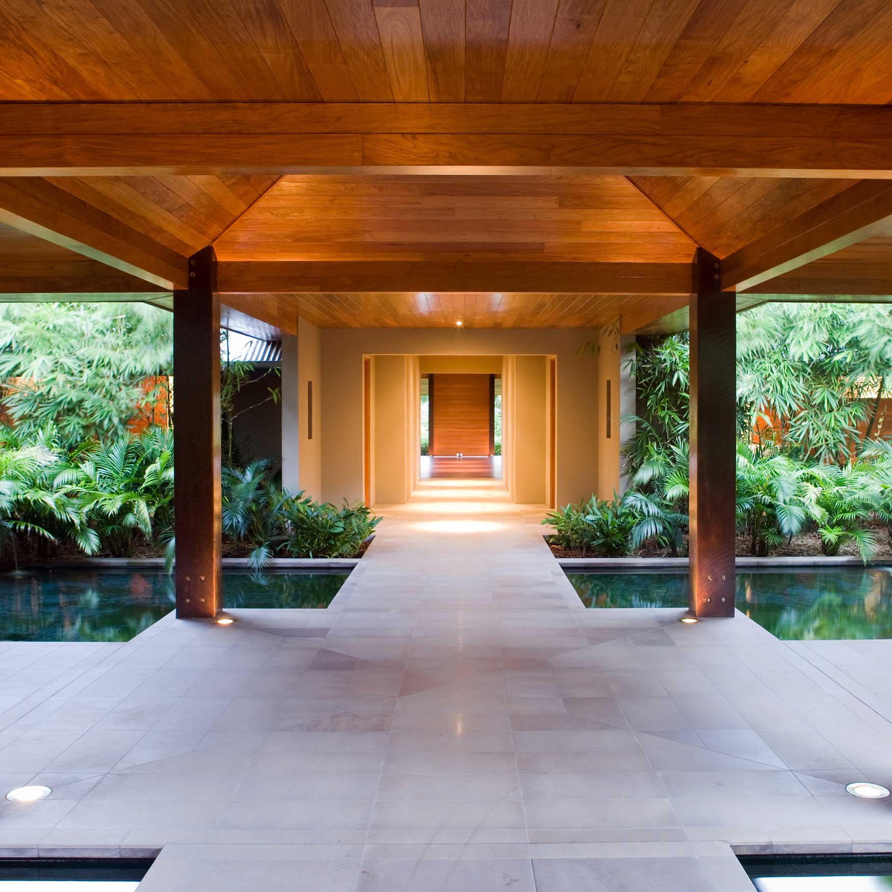 A modern, luxurious hallway featuring wooden beams and a polished stone floor, flanked by lush greenery. The space has soft lighting and a serene atmosphere, leading to an outdoor area with a reflecting pool.