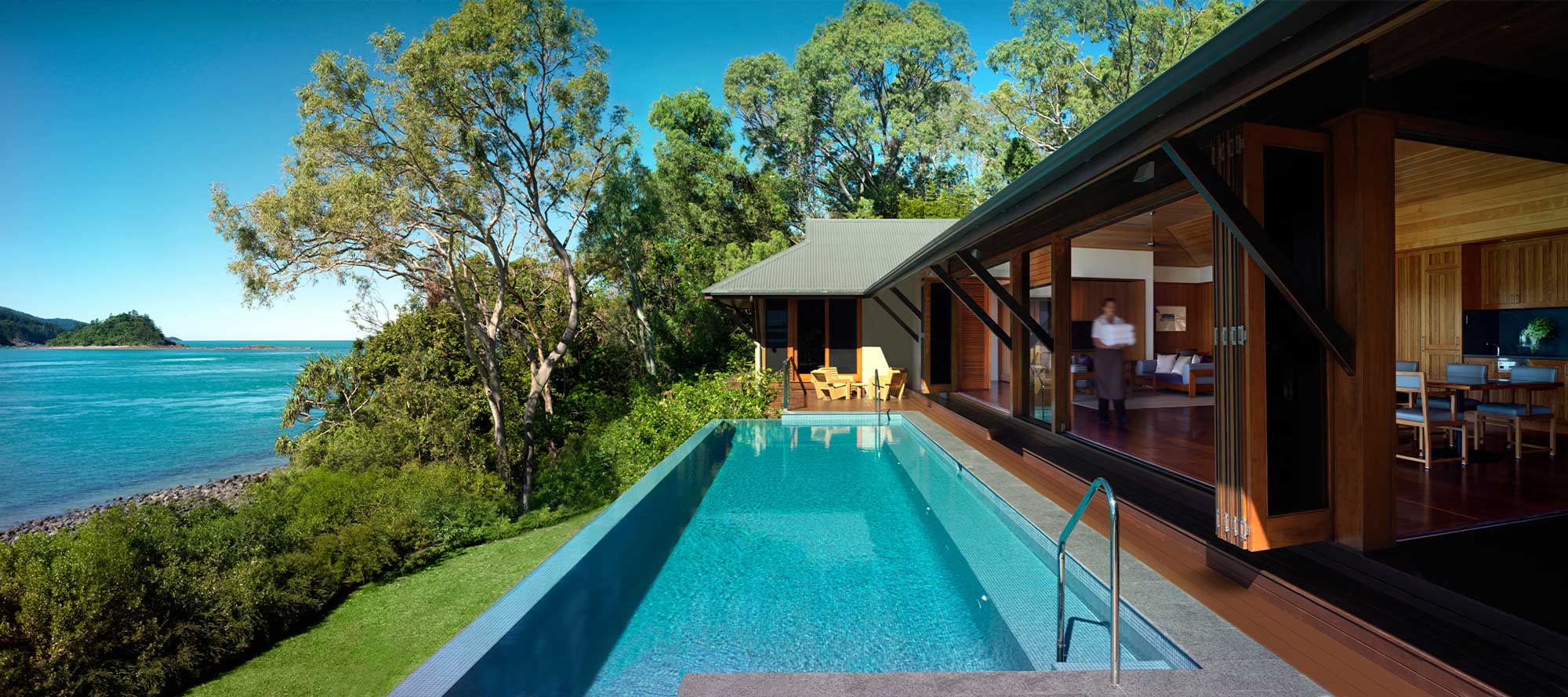 A modern home with large glass walls opens to an infinity pool, overlooking a serene coastal landscape. Lush greenery surrounds the property, and the ocean sparkles in the sunlight.
