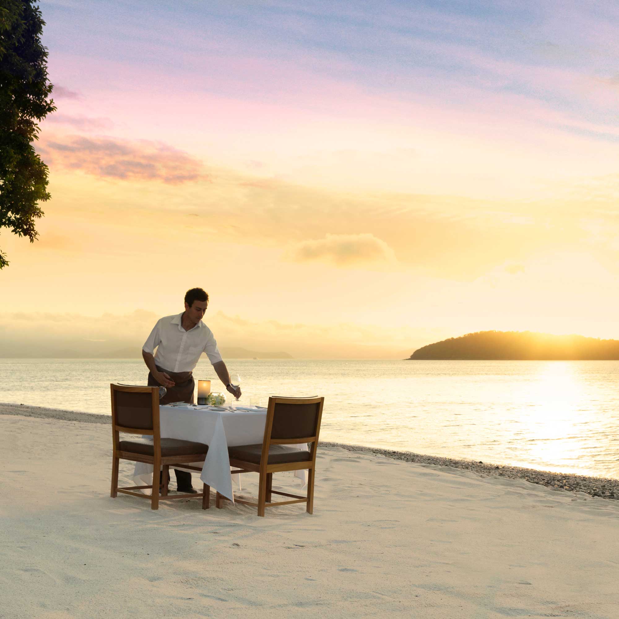 A waiter sets a table for two on a sandy beach during sunset, with calm waters and a distant island visible.