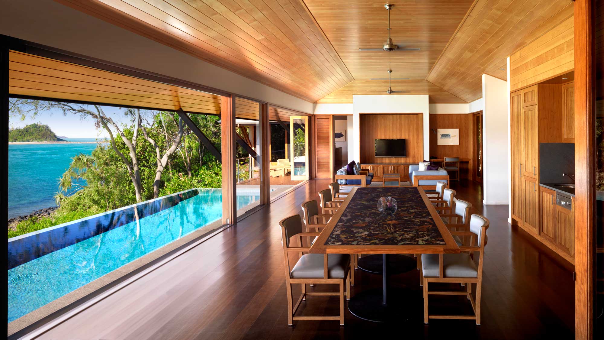 A spacious dining area with a long wooden table overlooks an infinity pool and scenic coastal views. Large windows and wooden accents create a bright, airy atmosphere.