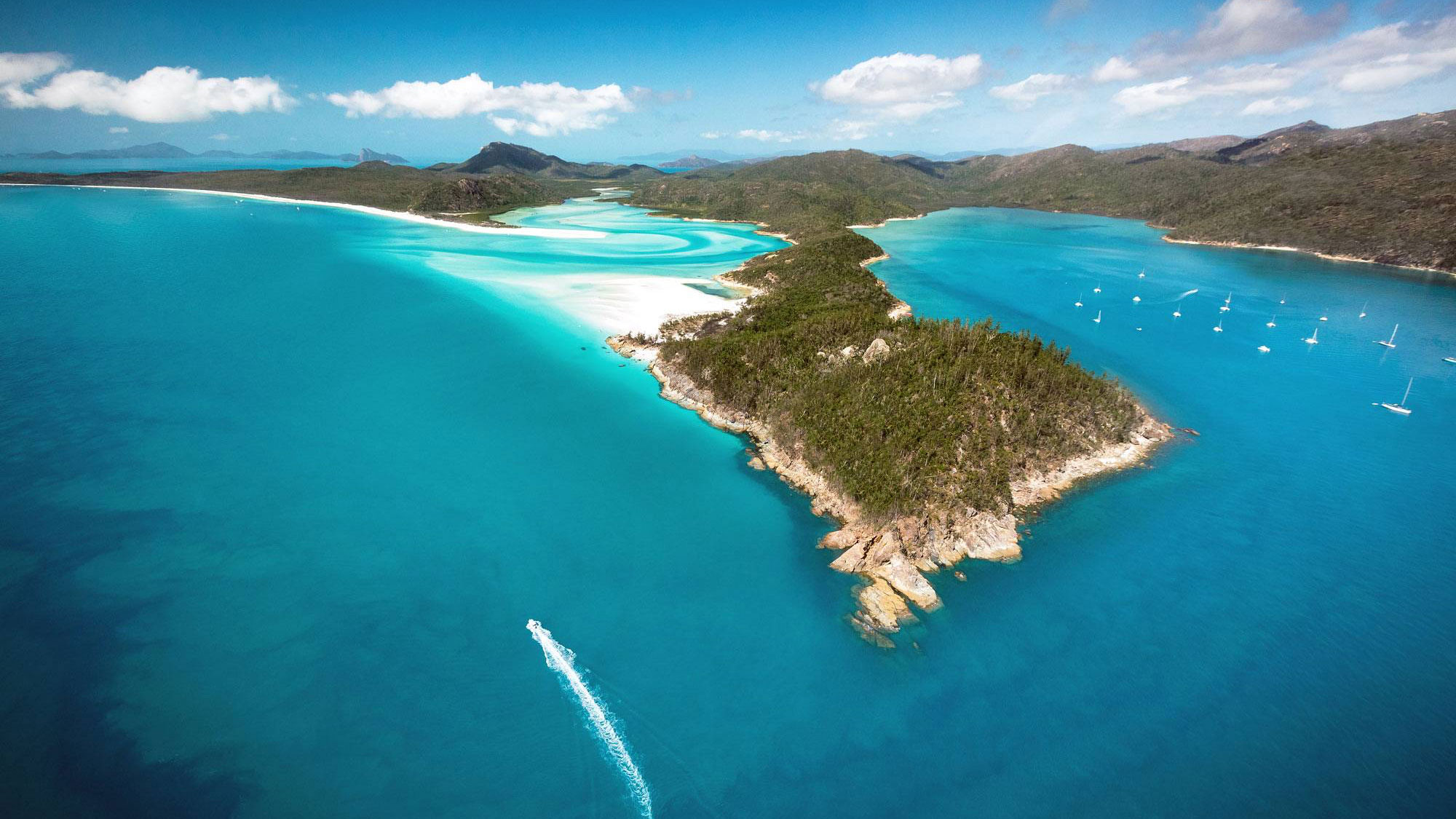 Aerial view of a tropical coastline featuring turquoise waters, white sandy beaches, and a lush green island with rocky outcrops. Sailboats are visible in the bay, and in the distance, rolling hills add to the scenic landscape.