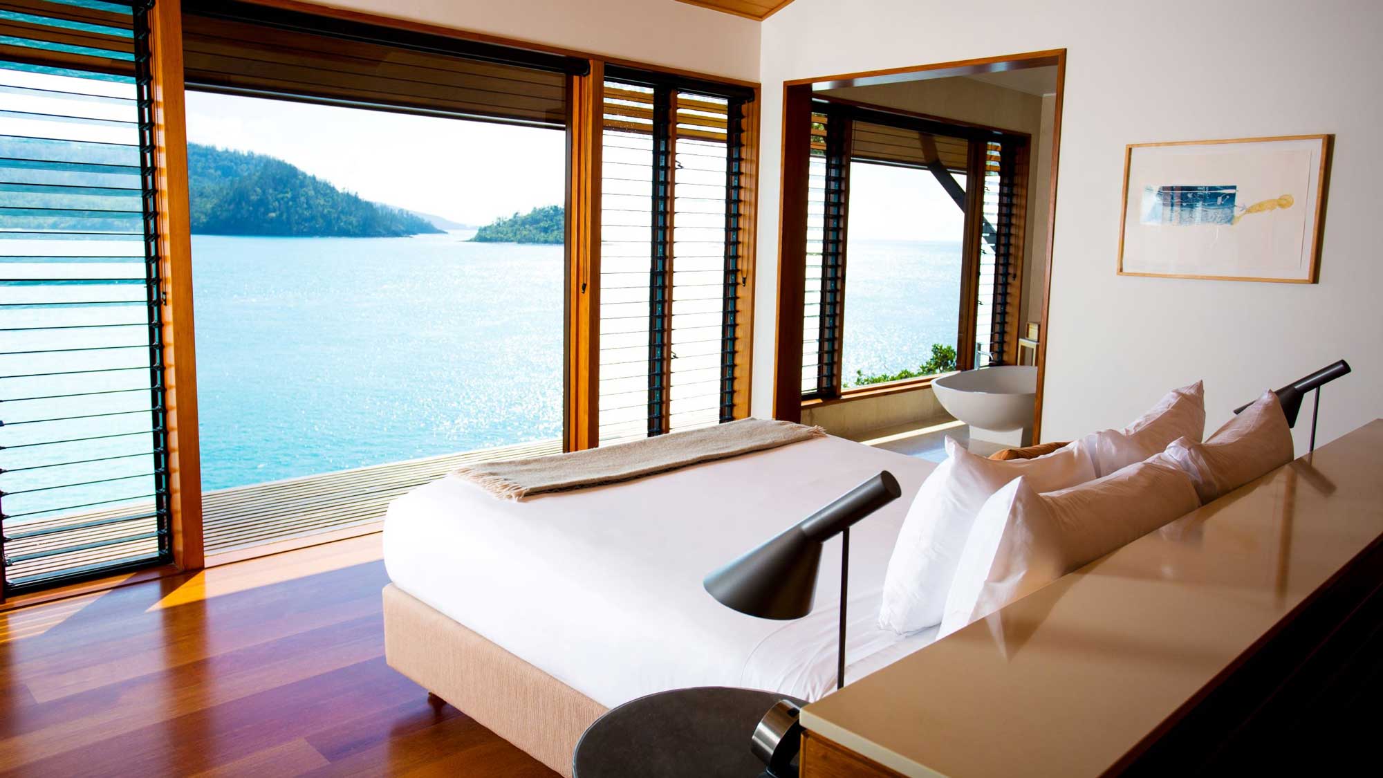 A modern bedroom with large windows overlooking a serene body of water and lush greenery. The room features a light-colored bed with white pillows, a sleek lamp on the side table, and a minimalist design that complements the scenic view.