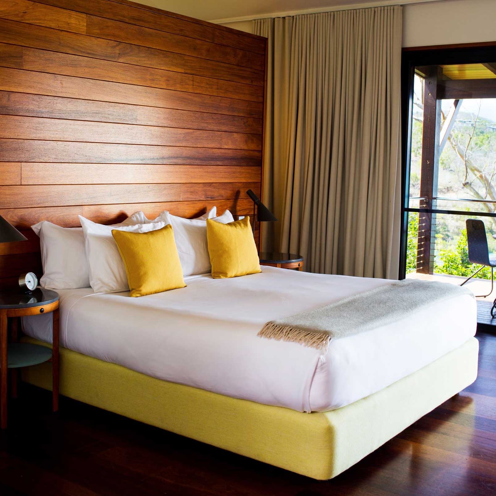 A cozy bedroom featuring a plush king-sized bed with white linens and yellow accent pillows. The wooden accent wall enhances the warm atmosphere, while large windows offer a view of the outdoors. A bedside table with a lamp and a light throw blanket add to the inviting decor.