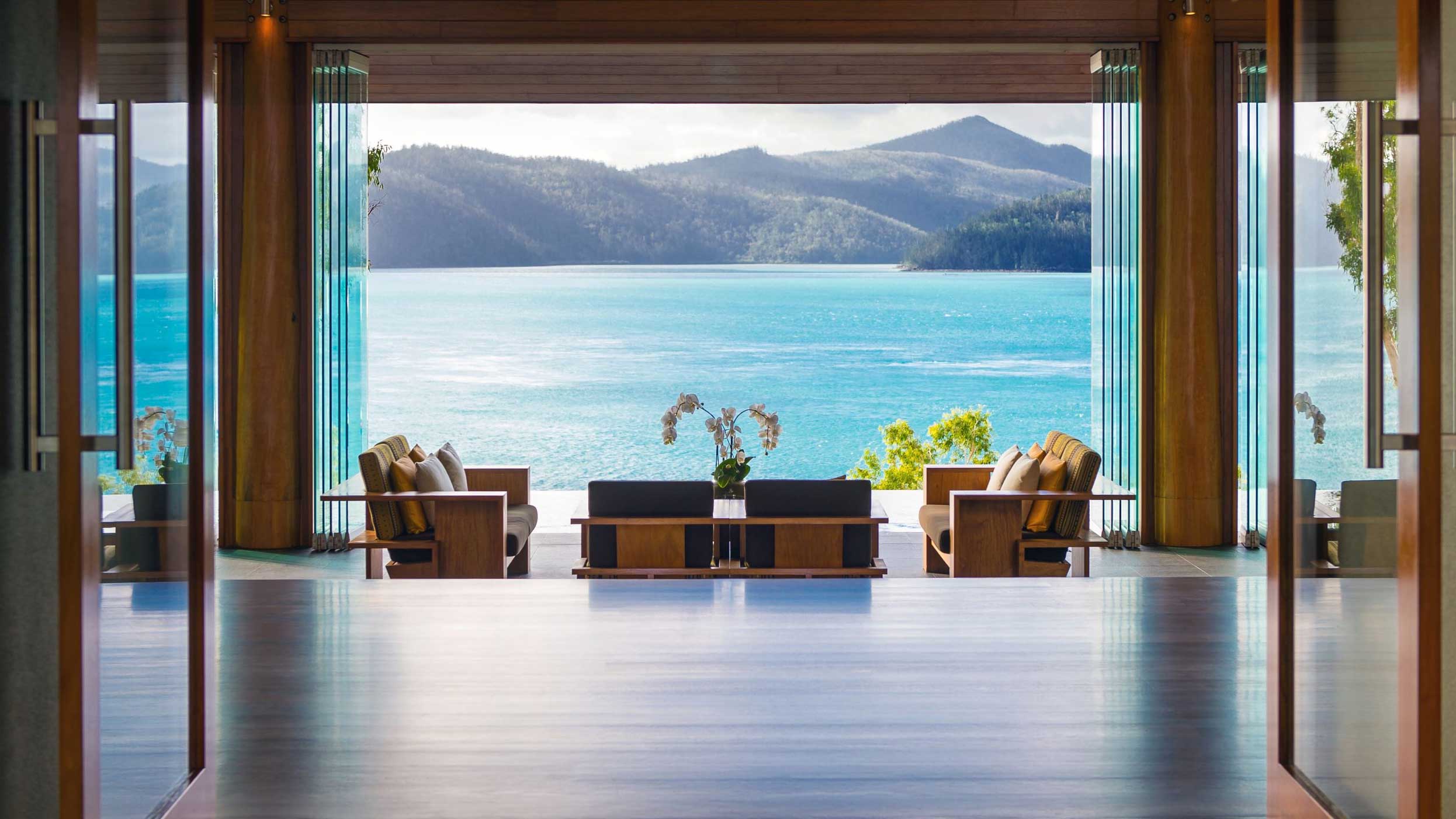 A modern interior featuring stylish furniture with a stunning view of a turquoise ocean and mountains in the distance through large glass doors. Natural light fills the space, creating a serene atmosphere.