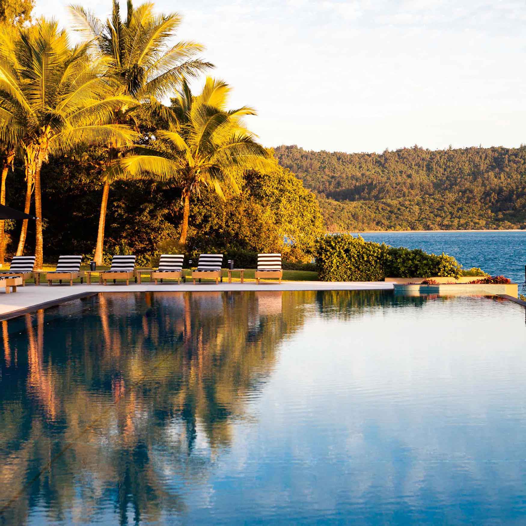 A serene swimming pool with lounge chairs beside it, surrounded by lush palm trees. The pool reflects the clear sky and nearby landscape, while a calm body of water and mountains are visible in the background.