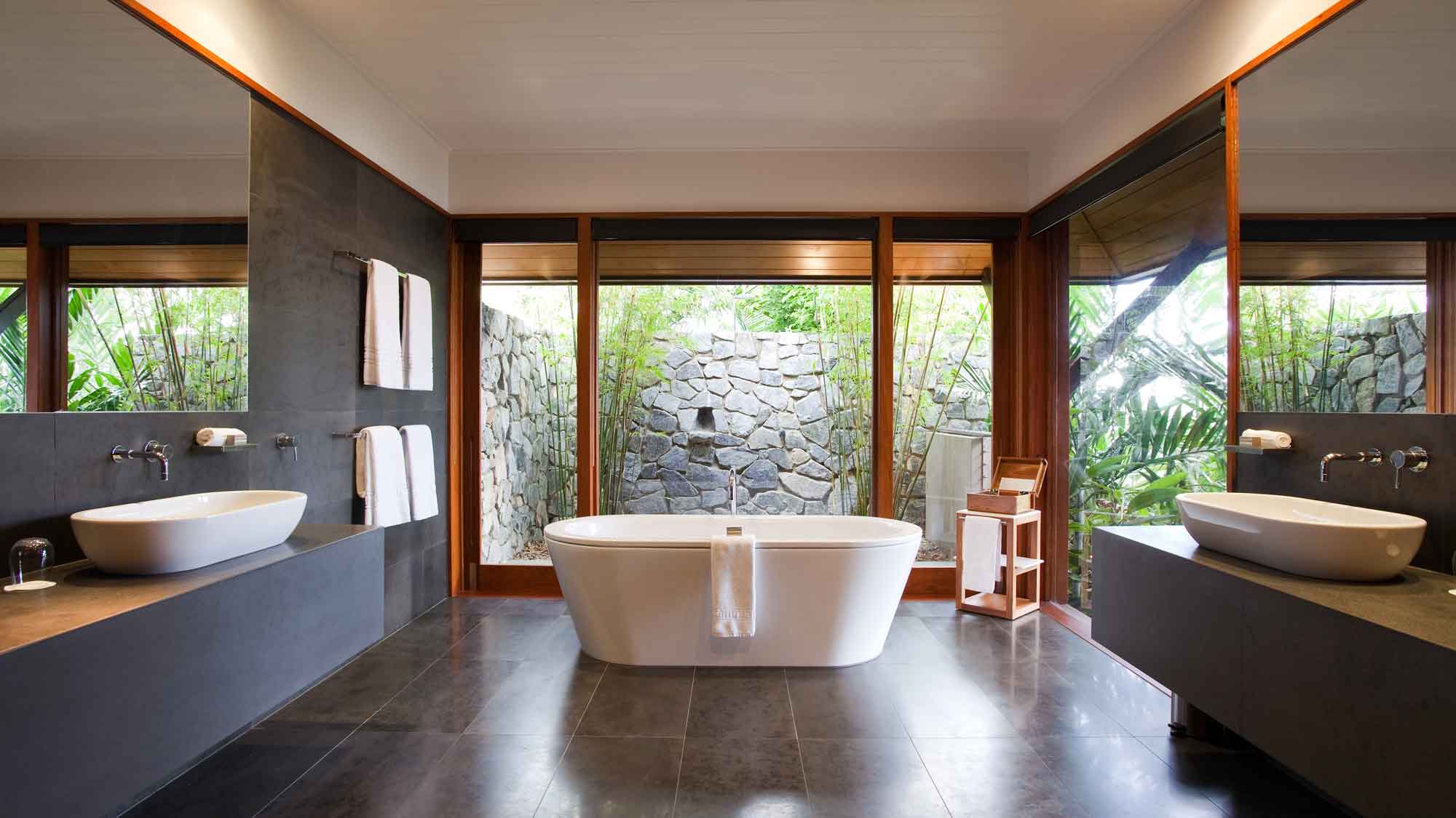 A modern bathroom featuring a freestanding bathtub, two sinks, and large windows with a view of a stone wall and greenery. The space has sleek, dark tiles and wooden accents. Towels hang neatly by the sink.