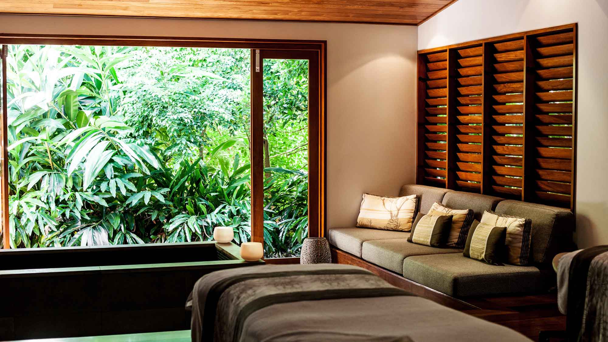 A serene spa room featuring a comfortable seating area with cushions, soft lighting, and a large window showcasing lush greenery outside.