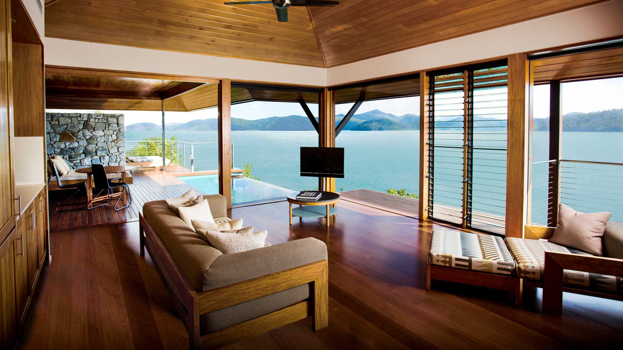 A modern living room with large windows offering a panoramic view of a tranquil lake and mountains. The room features wooden accents, a comfortable couch, a television, and a lounge area with a plush chair. Natural light floods the space, creating a warm atmosphere.