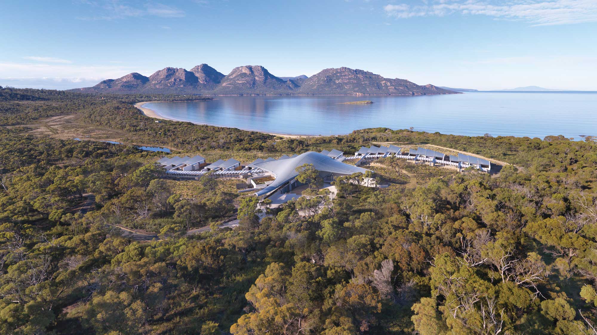 Aerial view of a modern resort nestled among trees near a calm shoreline, with mountains in the background and clear blue skies.