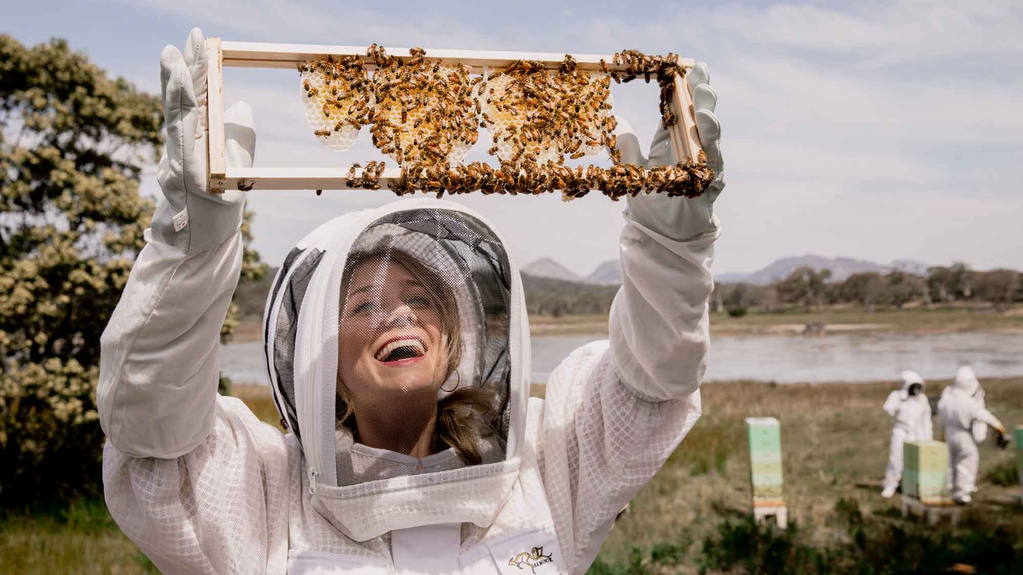 A smiling woman in a beekeeping suit holds up a frame filled with bees, showcasing her enthusiasm. In the background, other beekeepers work near hives in a natural setting.