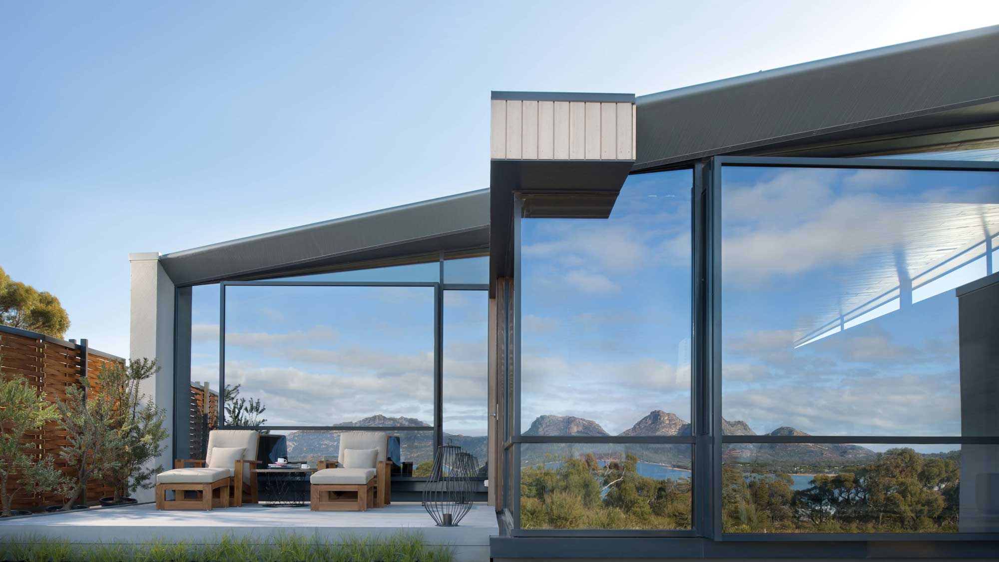 Modern architectural design featuring a patio with two lounge chairs. Large windows reflect a cloudy sky and showcase distant mountains and water views. Lush greenery surrounds the outdoor area.