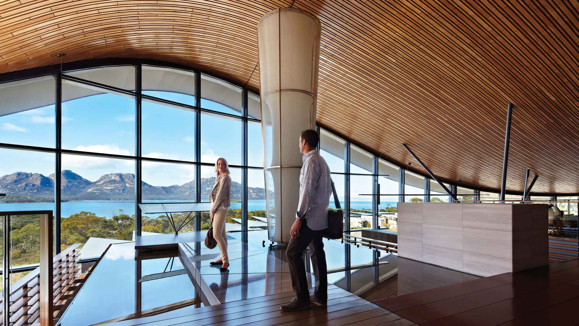 A modern interior space with a wooden ceiling, featuring large glass windows that showcase a scenic view of mountains and water. Two people, a man and a woman, are standing inside, with the woman oriented towards the view.