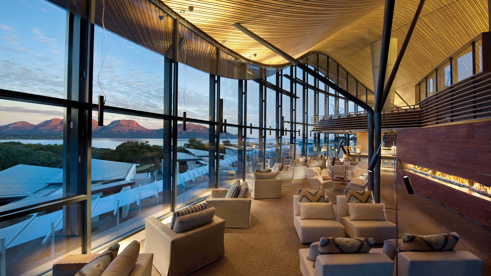 A modern lounge with large glass windows showcasing a scenic view of mountains and water. Stylish white sofas and armchairs create a welcoming atmosphere, illuminated by warm lighting.