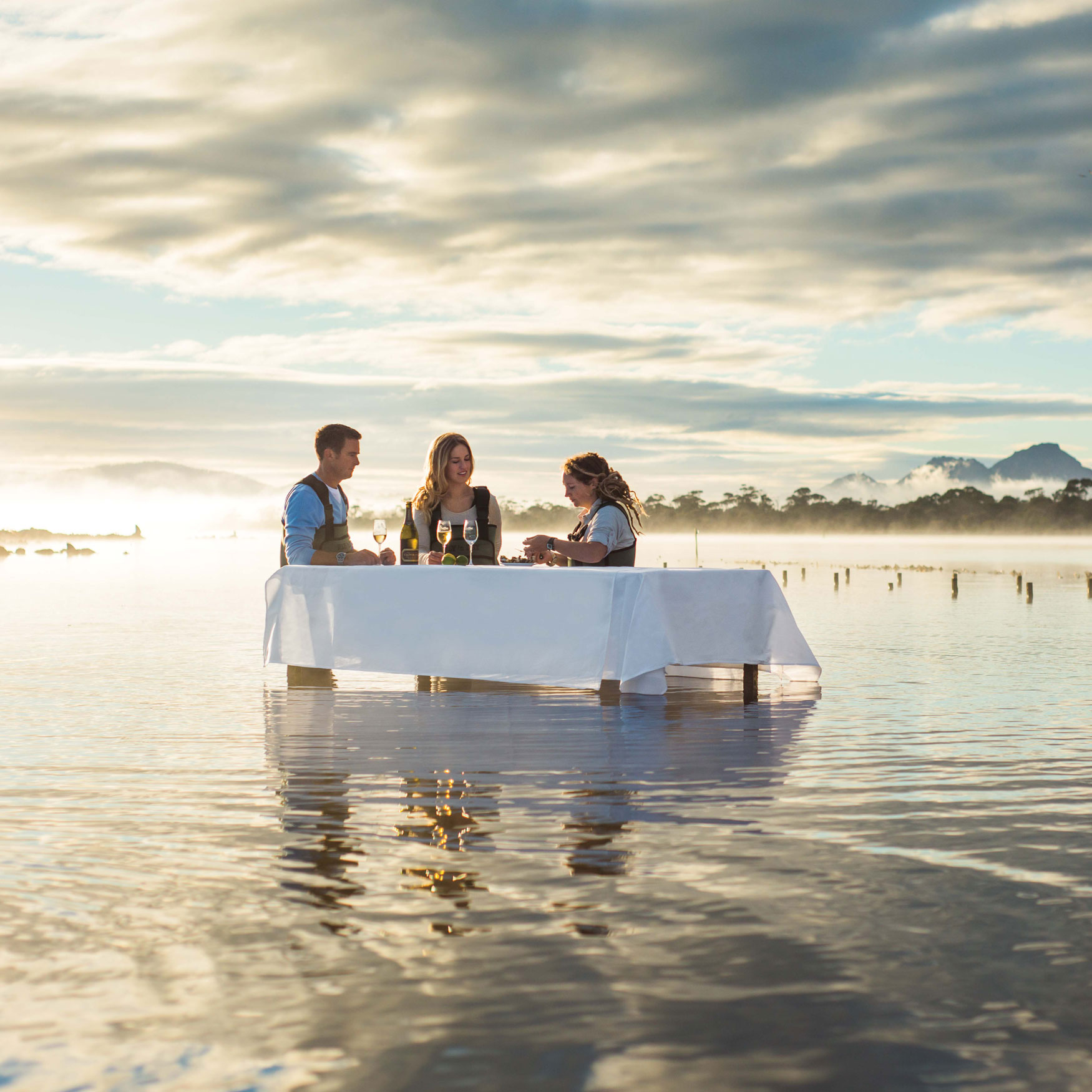 Three people sit at a white table set in shallow water, surrounded by misty scenery and mountains in the background. They enjoy drinks while engaged in conversation under a cloudy sky.