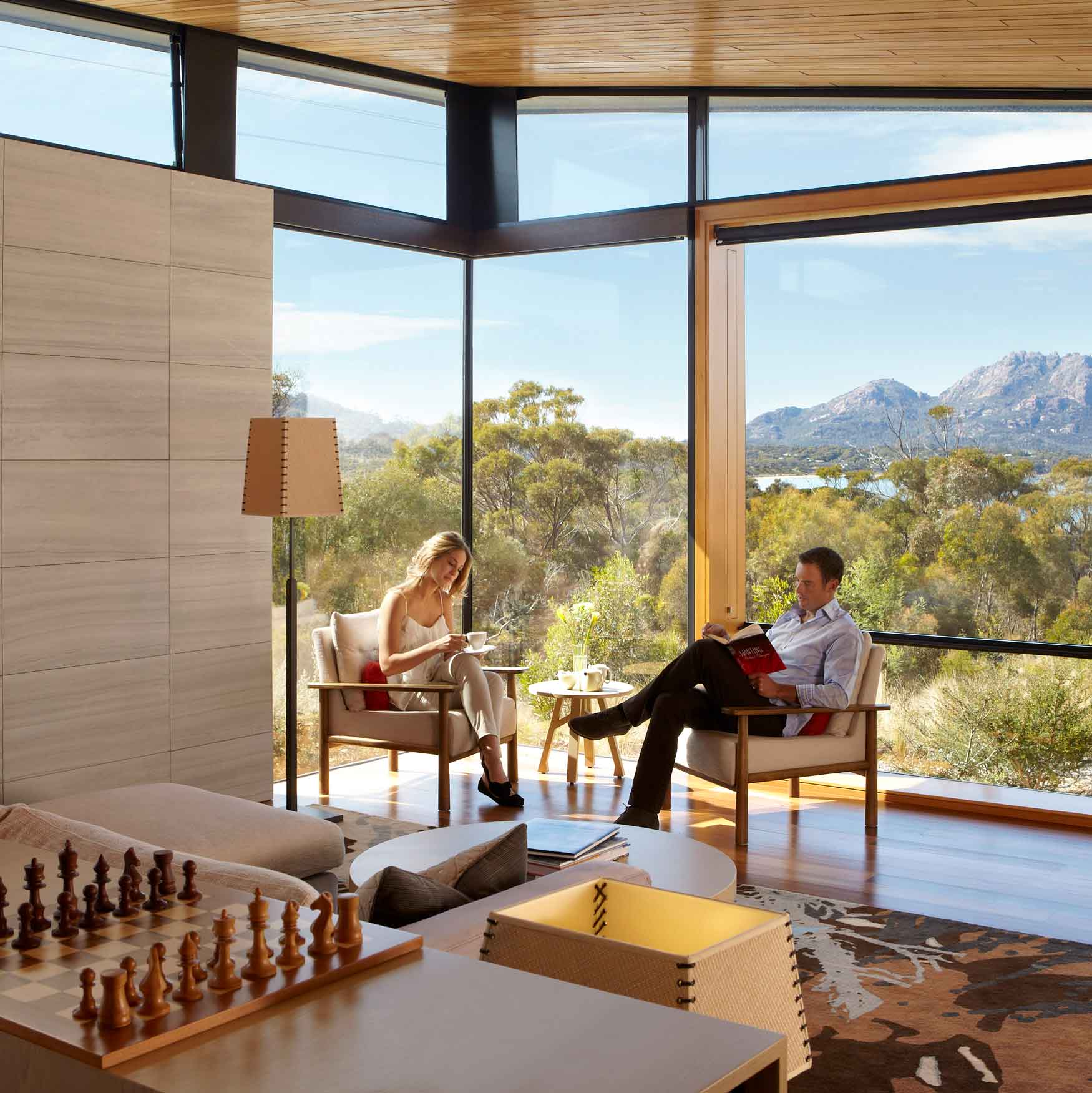 A modern living room features large windows with a scenic view of mountains and trees. A woman sits at a table reading, while a man relaxes in an armchair, looking at a magazine. A chess set is visible on a nearby table.