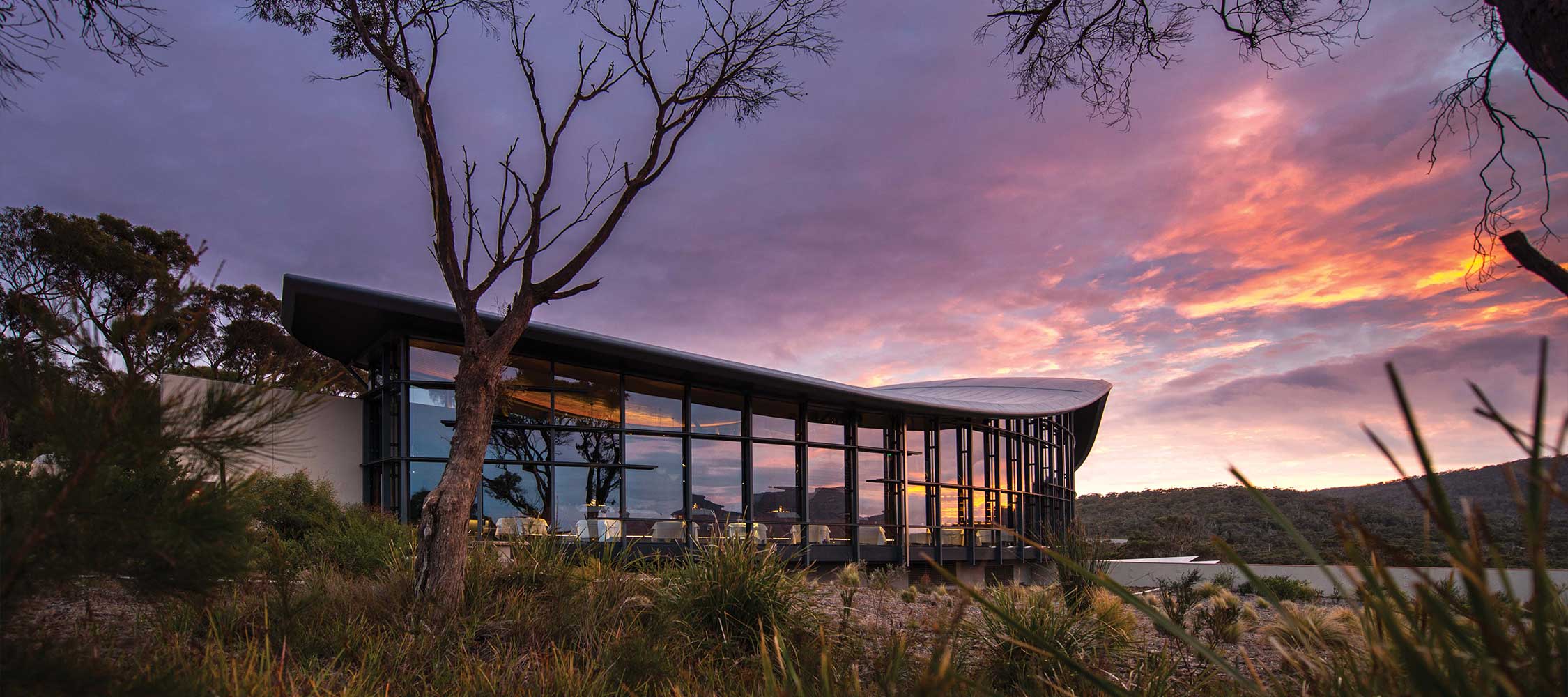 A modern architectural building with large glass windows reflects a colorful sunset, surrounded by trees and lush vegetation. The sky displays vibrant shades of purple and orange, enhancing the serene landscape.