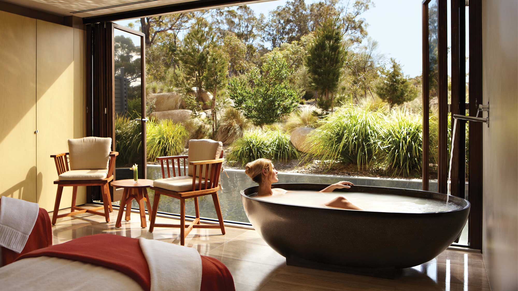 A relaxing spa scene featuring a person soaking in a round black bathtub. Sunlight streams in through large glass doors, revealing a serene outdoor garden with lush greenery. Two wooden chairs and a small table are positioned nearby, enhancing the tranquil atmosphere.