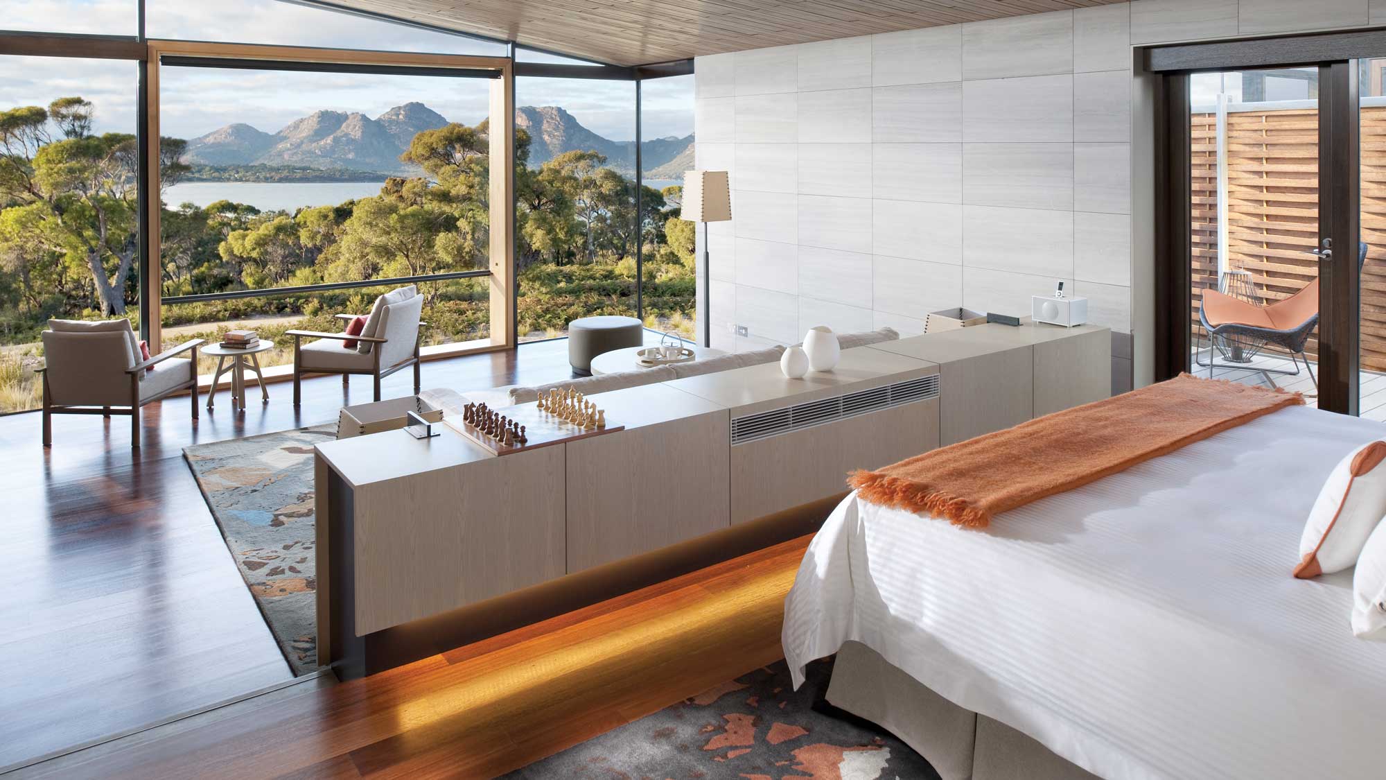 A modern bedroom with a large window showcasing a scenic view of mountains and a lake. The room features wooden flooring, a plush bed with an orange throw, and a stylish seating area. A chess set is displayed on a sleek, contemporary console.