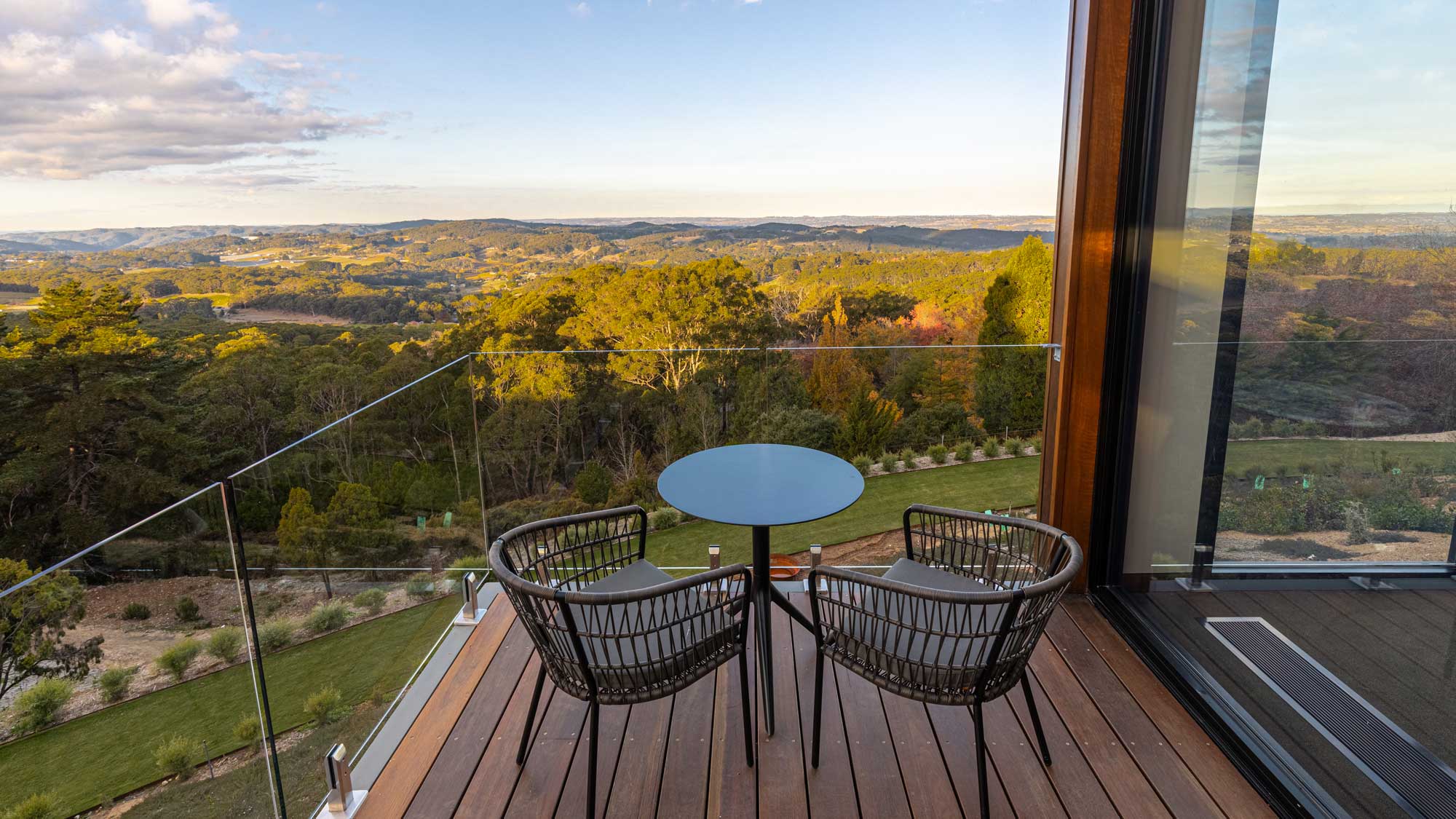 A balcony with two chairs and a small table overlooks scenic hills and valleys with lush greenery under a clear sky.